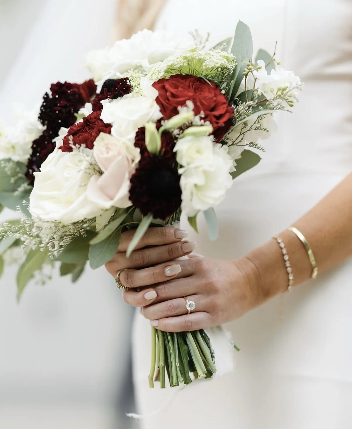 A bride holding a bridal bouquet of red, white, and blush flowers with green foliage, wearing a gold bracelet and a diamond engagement ring.