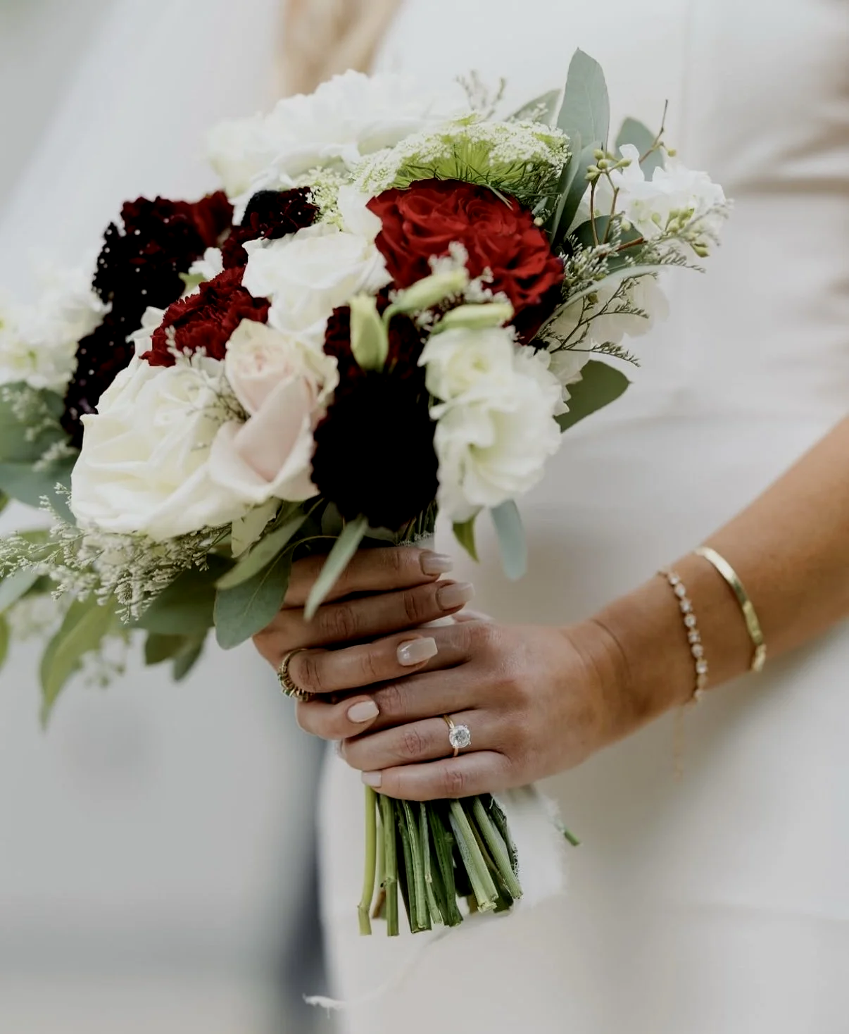 Close-up of a bride holding a bouquet of red, white, and cream flowers with green leaves, wearing a diamond engagement ring, a thin bracelet, and a gold bangle.