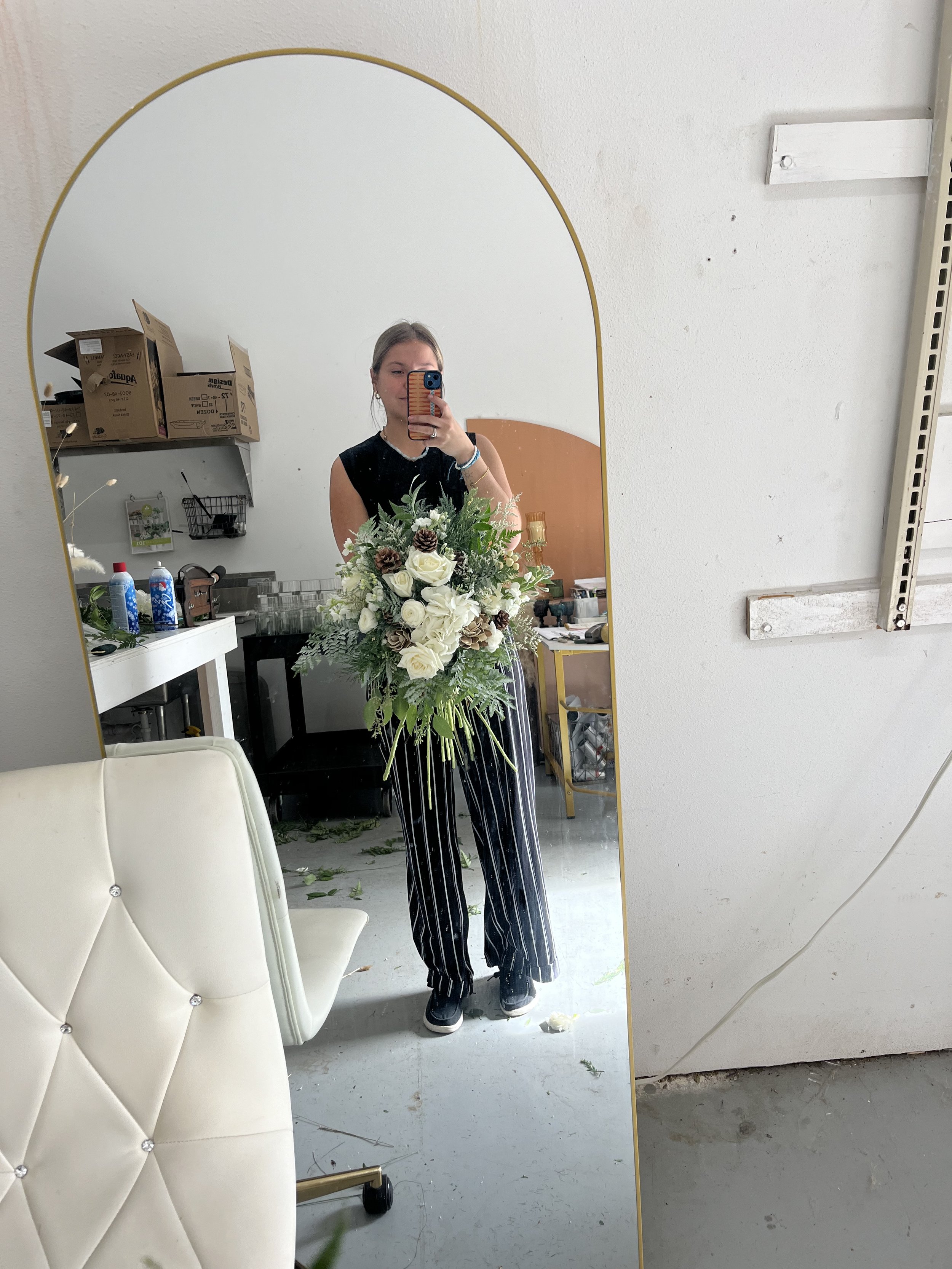 A woman taking a selfie in a tall, arched mirror, holding a large bouquet of white roses and pinecones. She is dressed in a black sleeveless top, black and white striped pants, and sneakers. The background shows a cluttered room with boxes, a white table, and some drawers.