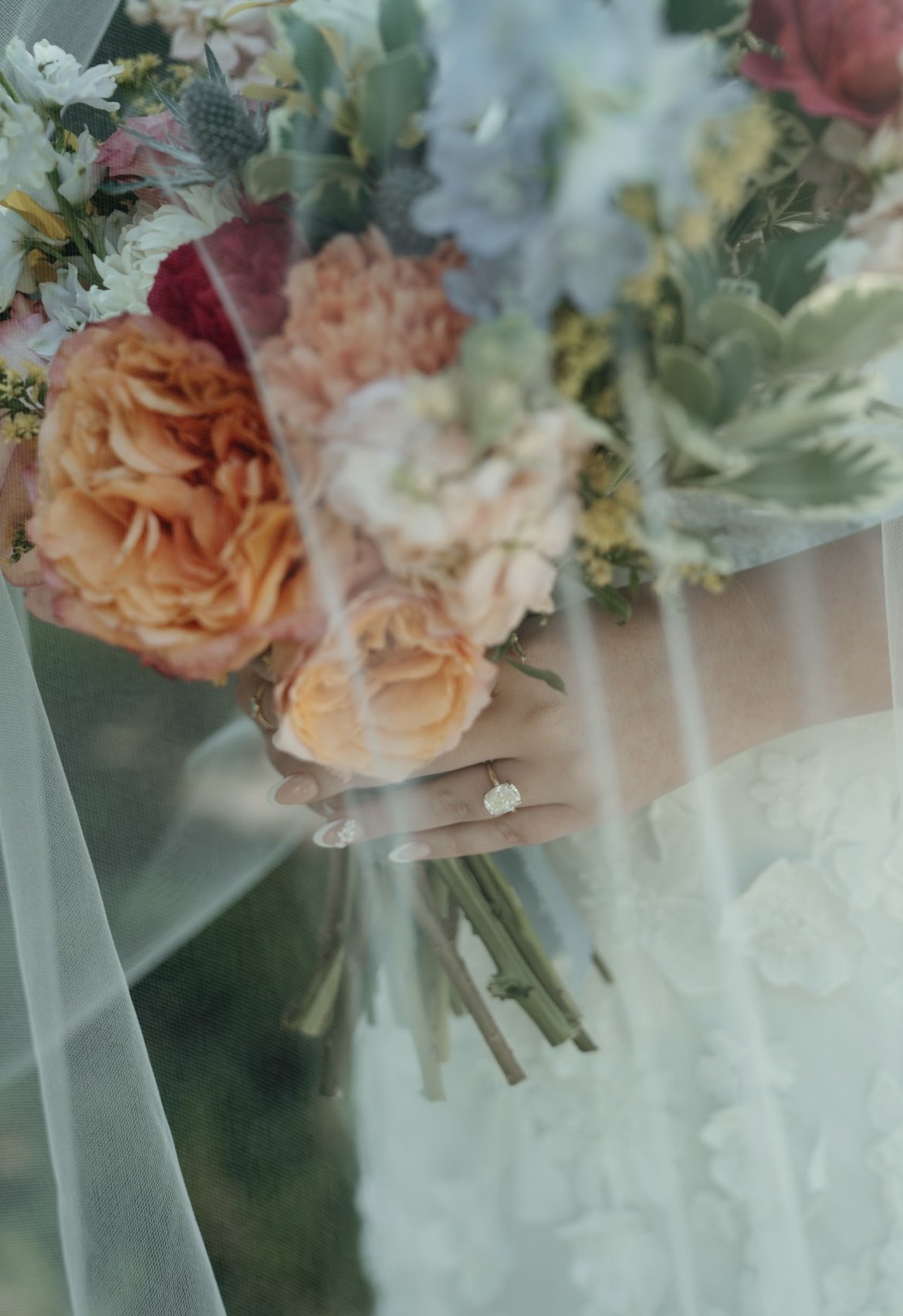 A close-up of a bride's hand with a glittering engagement ring holding a bouquet of orange, white, and pink flowers seen through a sheer veil.