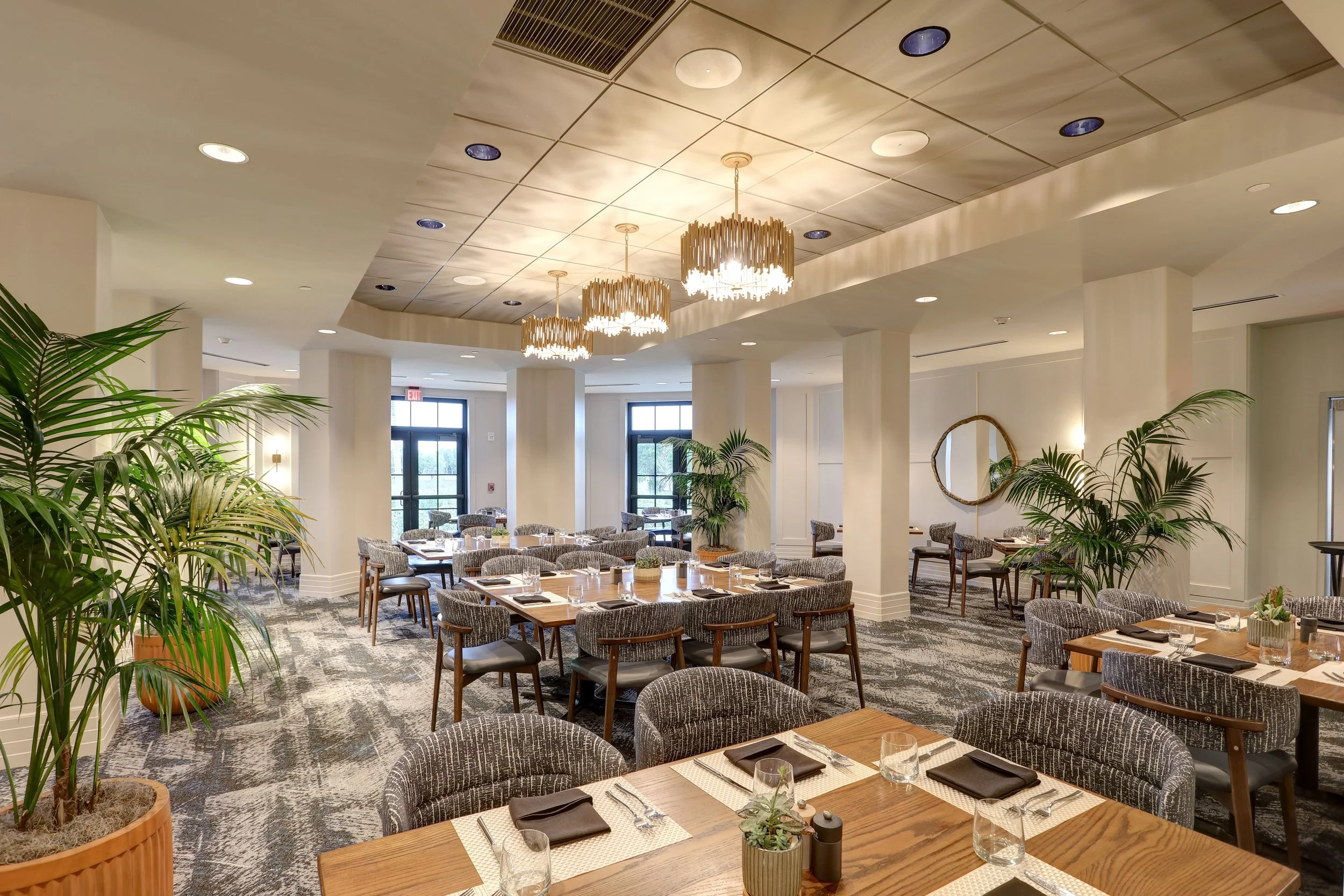 Elegant restaurant interior with round tables set for dining, gray upholstered chairs, large potted plants, and modern gold chandeliers hanging from a patterned ceiling.