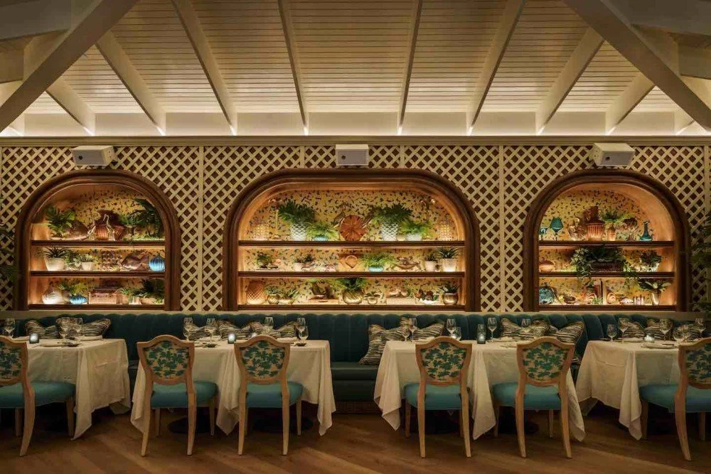 Elegant dining room with seven tables, each set with glasses and plates. There are eight chairs with teal cushions and floral upholstery around the tables. The background features three illuminated shelves with decorative plants and vases, set against a lattice wall under a sloped ceiling.