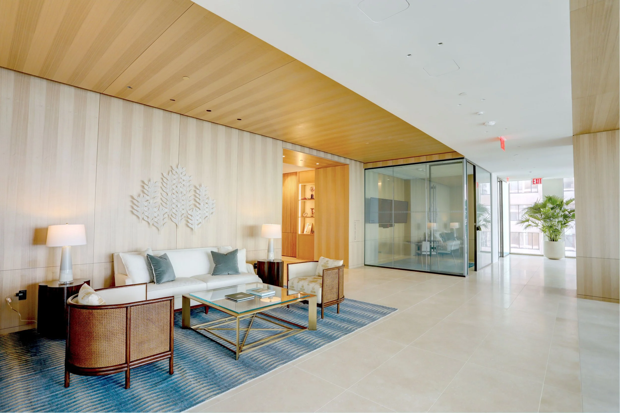 Modern hotel lobby with a white sofa, armchair, and coffee table on a blue striped rug, wooden panel walls with a metal art piece, glass-walled conference room, and large potted plant near glass doors.