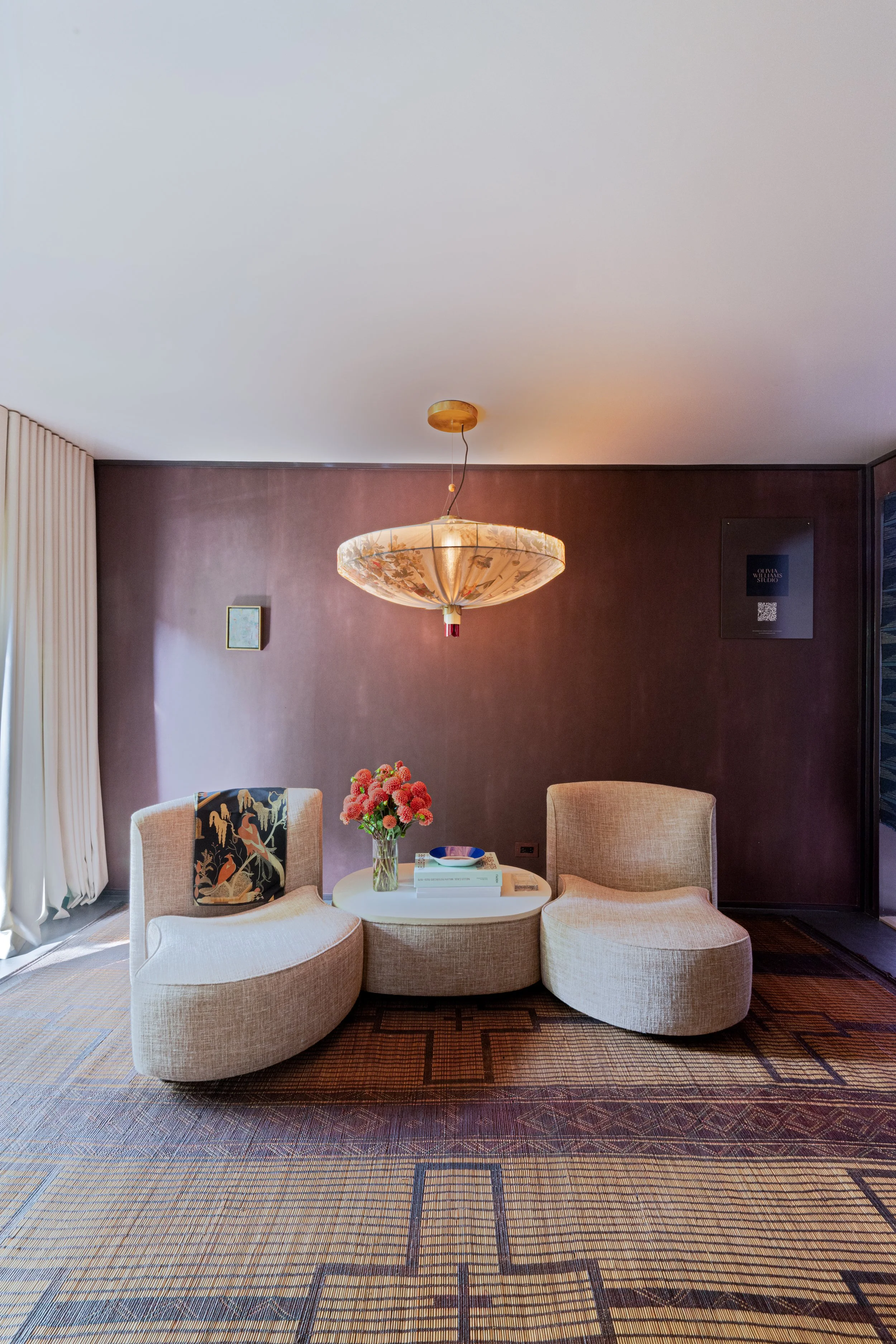 Modern hotel lobby with two beige curved chairs, a round white coffee table, a pink floral arrangement, a dark wood accent wall, and a large decorative ceiling light.
