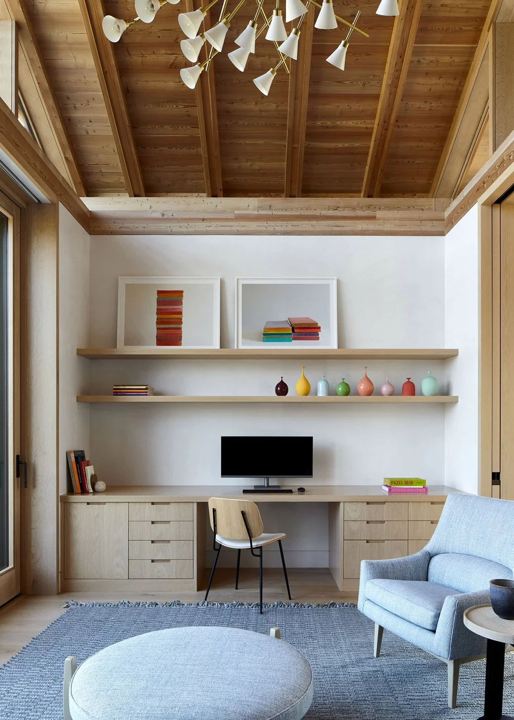 Living room with wood-paneled vaulted ceiling, white walls, built-in desk with a small computer monitor, shelves with colorful vases and books, armchair, ottoman, and large window.