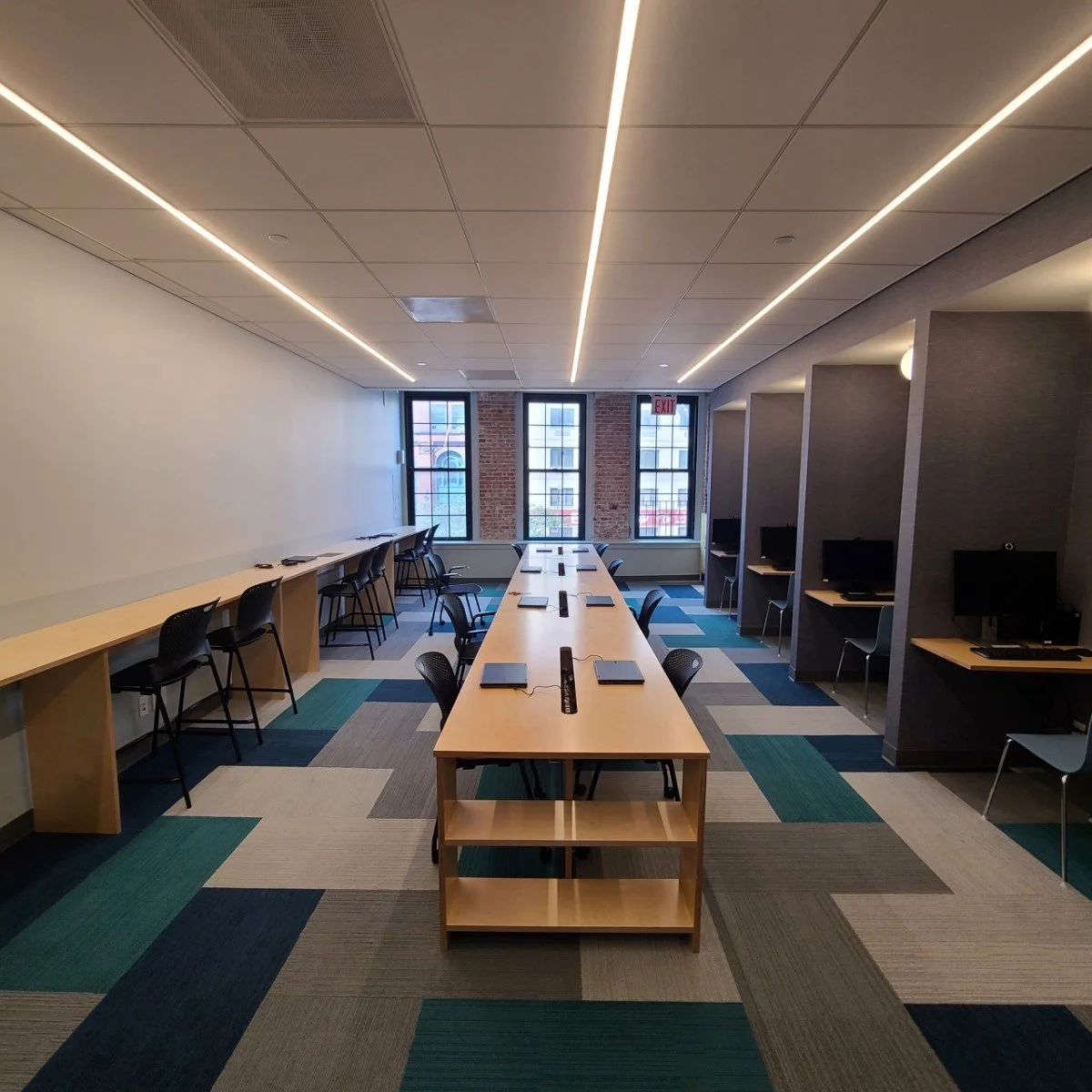 Modern computer lab or workspace with a long table and chairs, desktop computers in individual cubicles, large windows at the end, and a ceiling with integrated lighting.