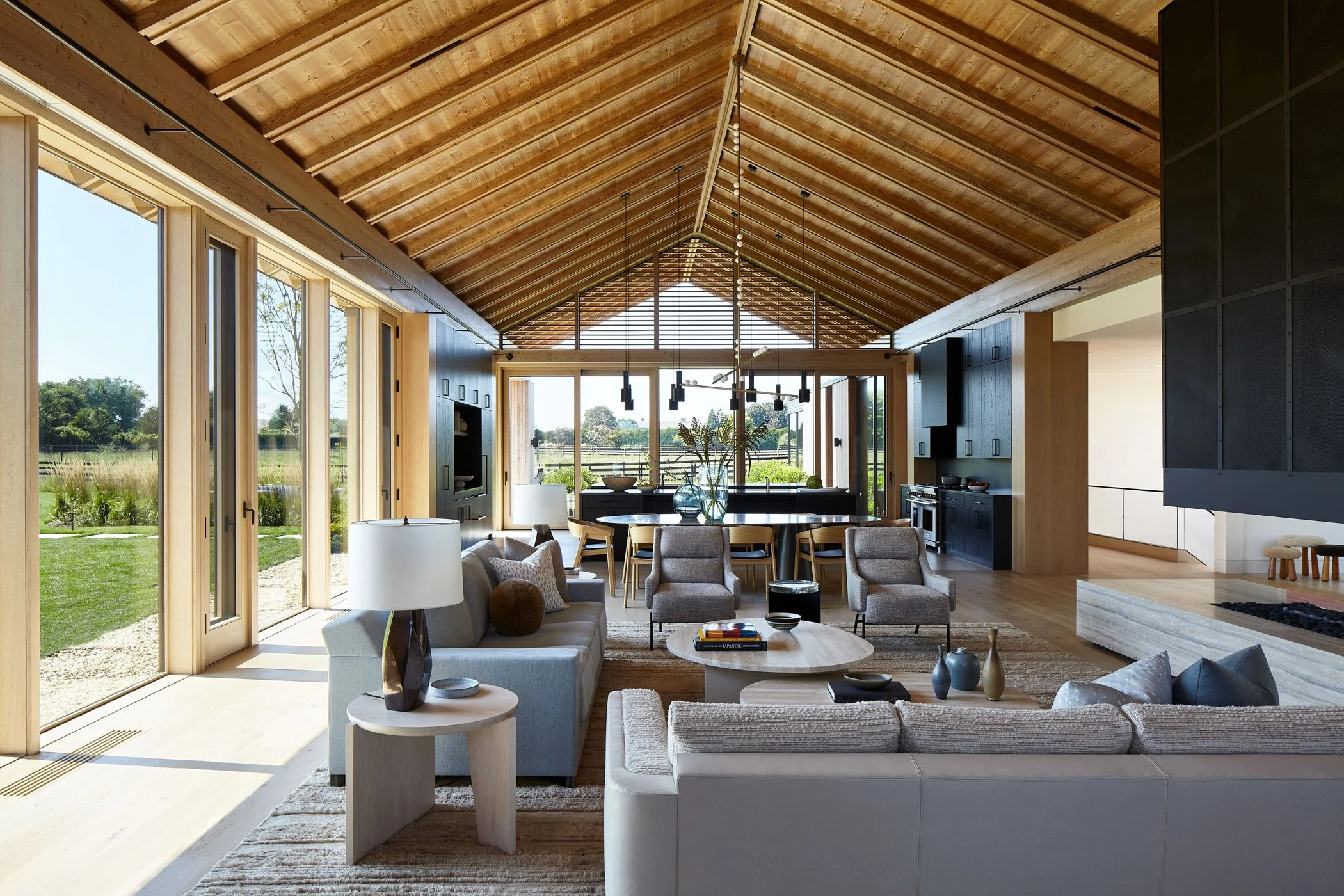 Spacious living room with wood vaulted ceiling, large glass doors opening to a garden, beige sofas, chairs, coffee tables, and a dining area with a black kitchen in the background.