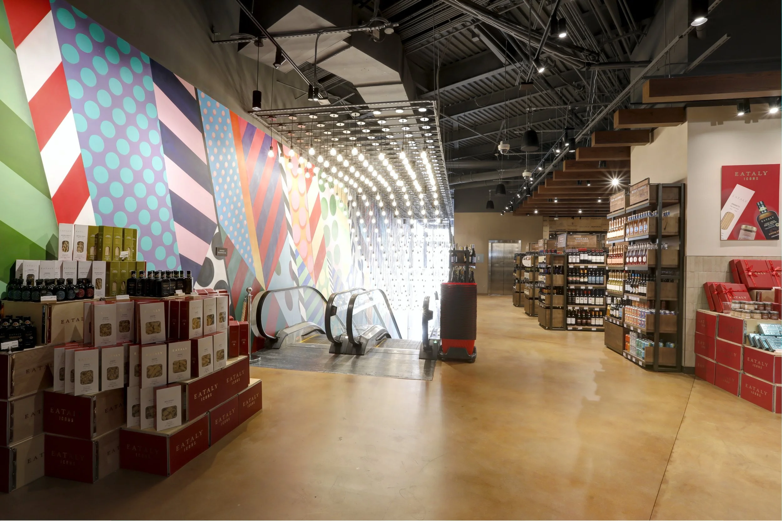 Interior of a retail store with colorful geometric mural wall, shelves stocked with various products, and escalators.