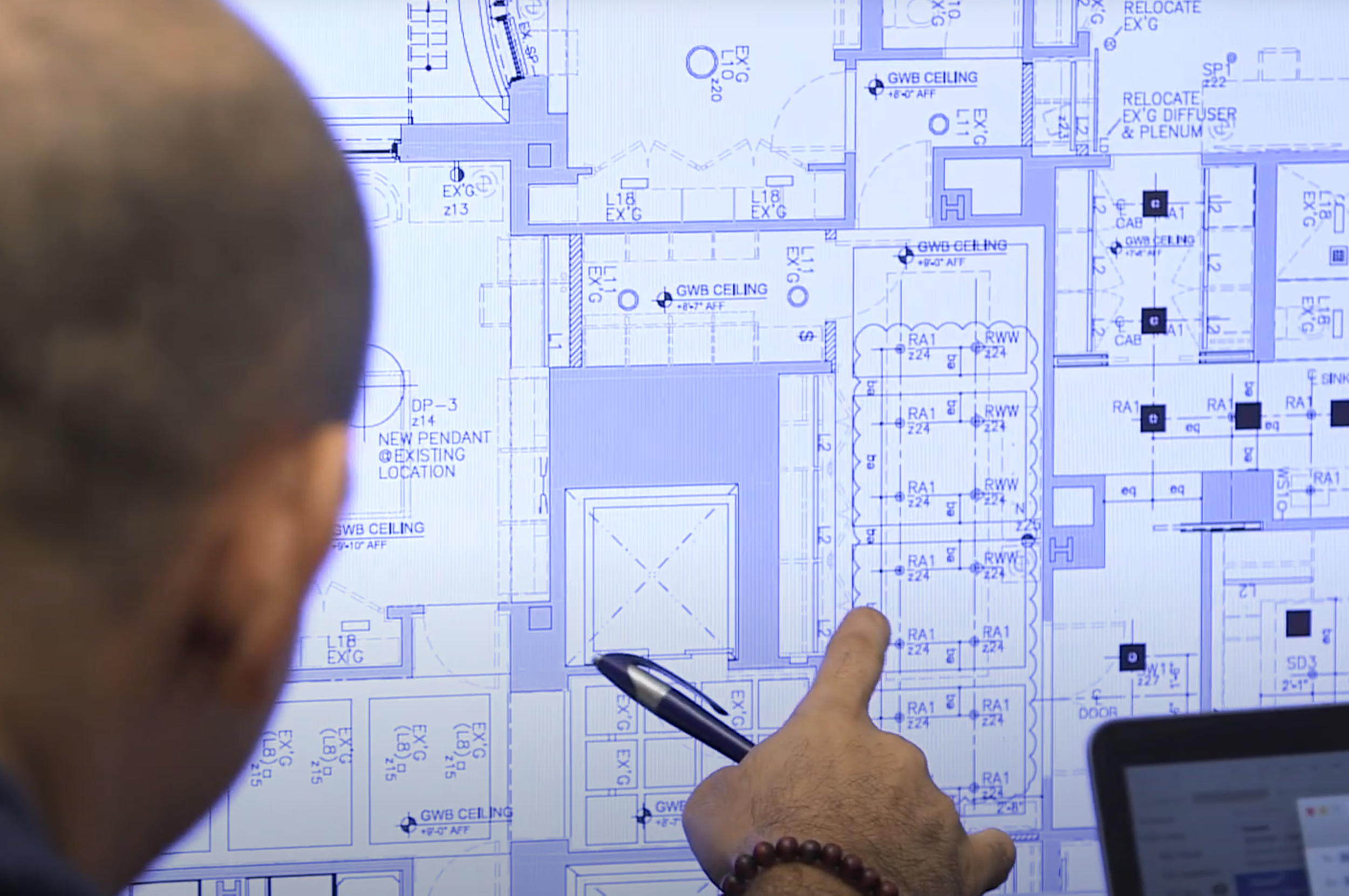 Person pointing at architectural blueprint on wall screens, showing building layout and electrical plans.