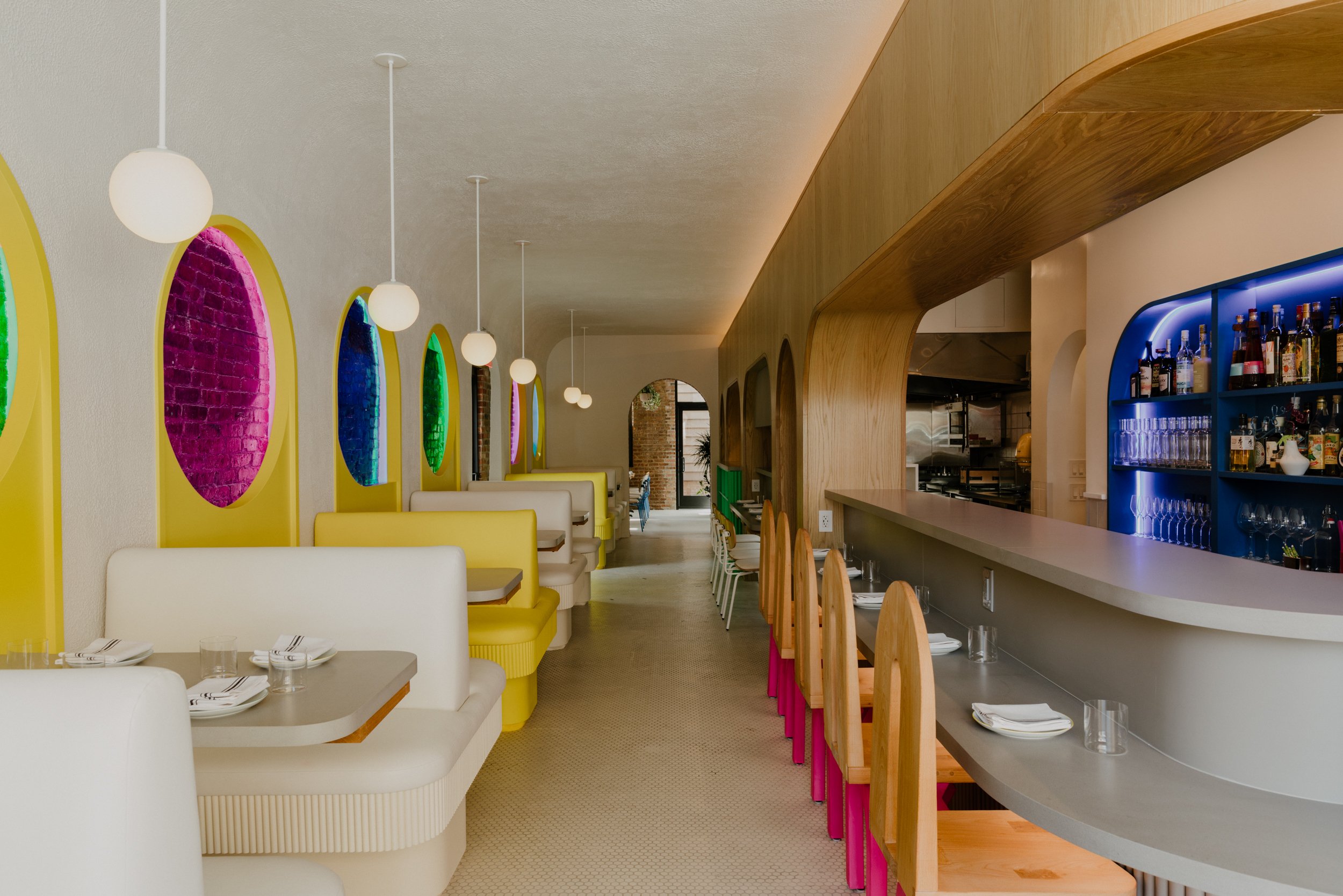 Modern restaurant interior with white and yellow booth seating on the left, colorful circular wall decorations, a bar counter on the right with wooden chairs, and hanging white pendant lights.