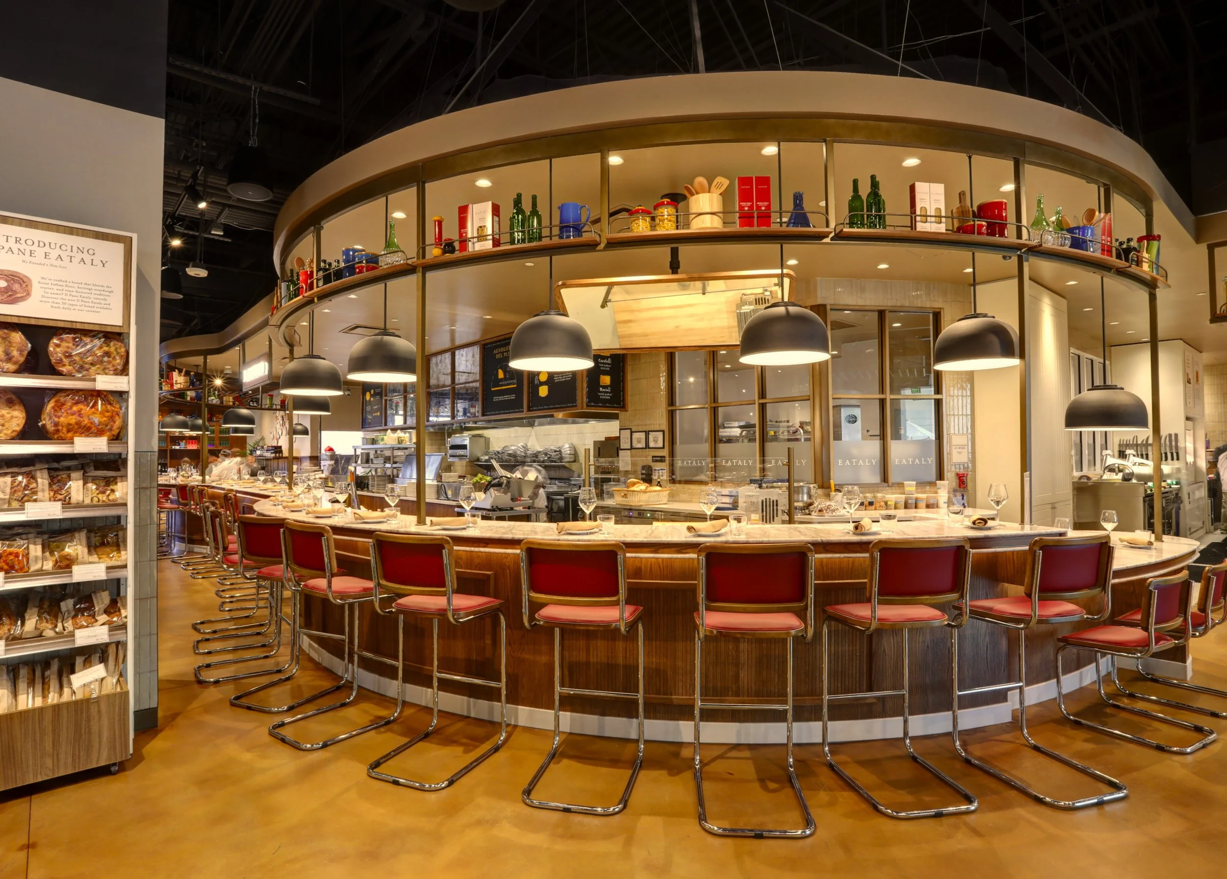 A circular bar in a restaurant with red chairs, black pendant lights, and shelves with bottles and kitchen items on the upper level.