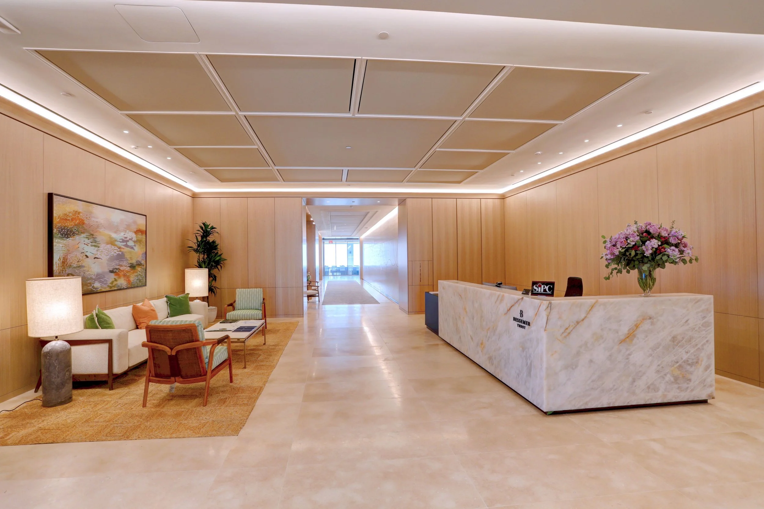 Hotel lobby with a reception desk on the right, a sitting area with sofas, chairs, and lamps on the left, and a hallway leading to a glass door at the back.