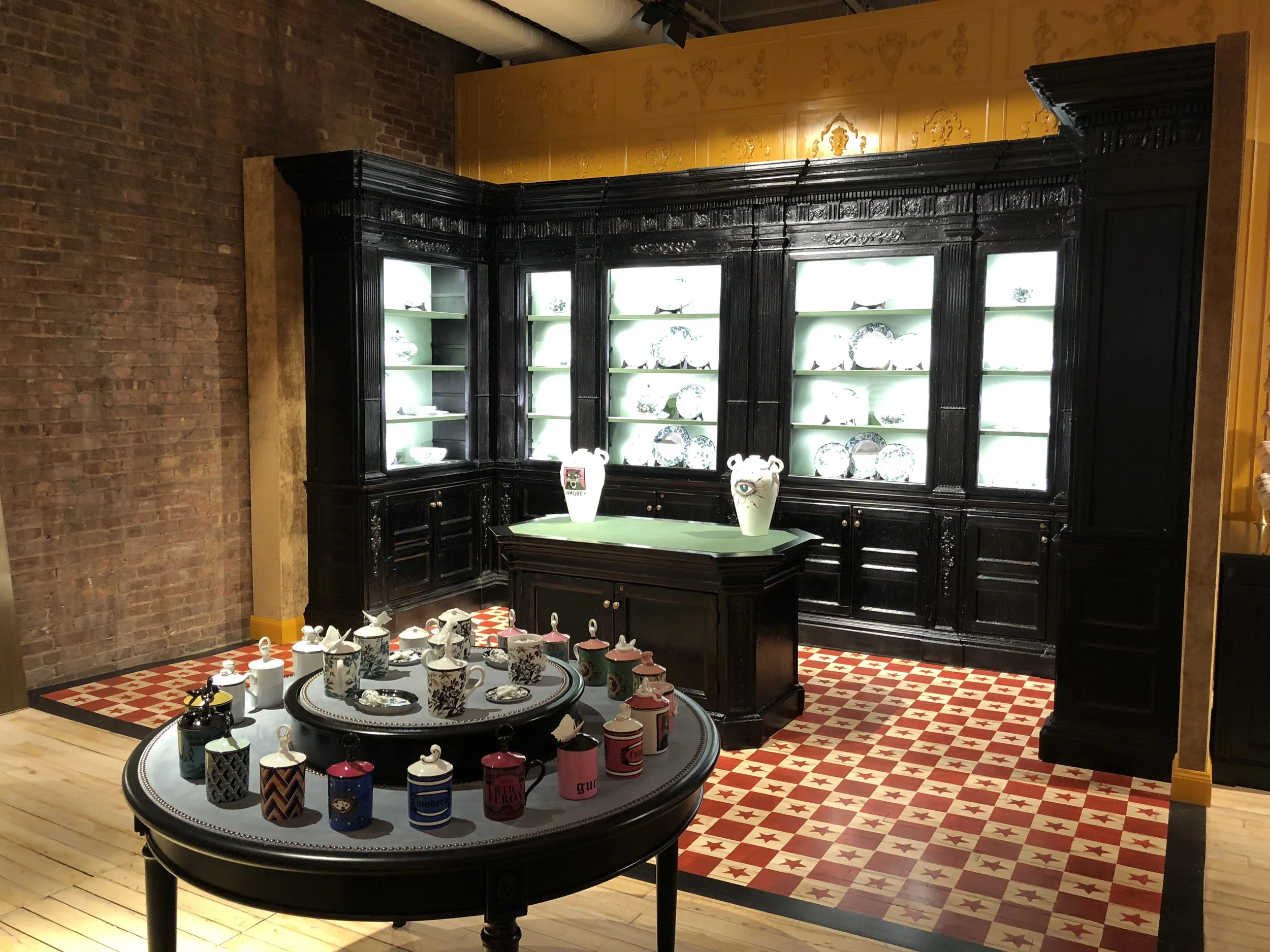 Display of decorative plates and cups in a gallery with black wooden shelving and a patterned tiled floor.