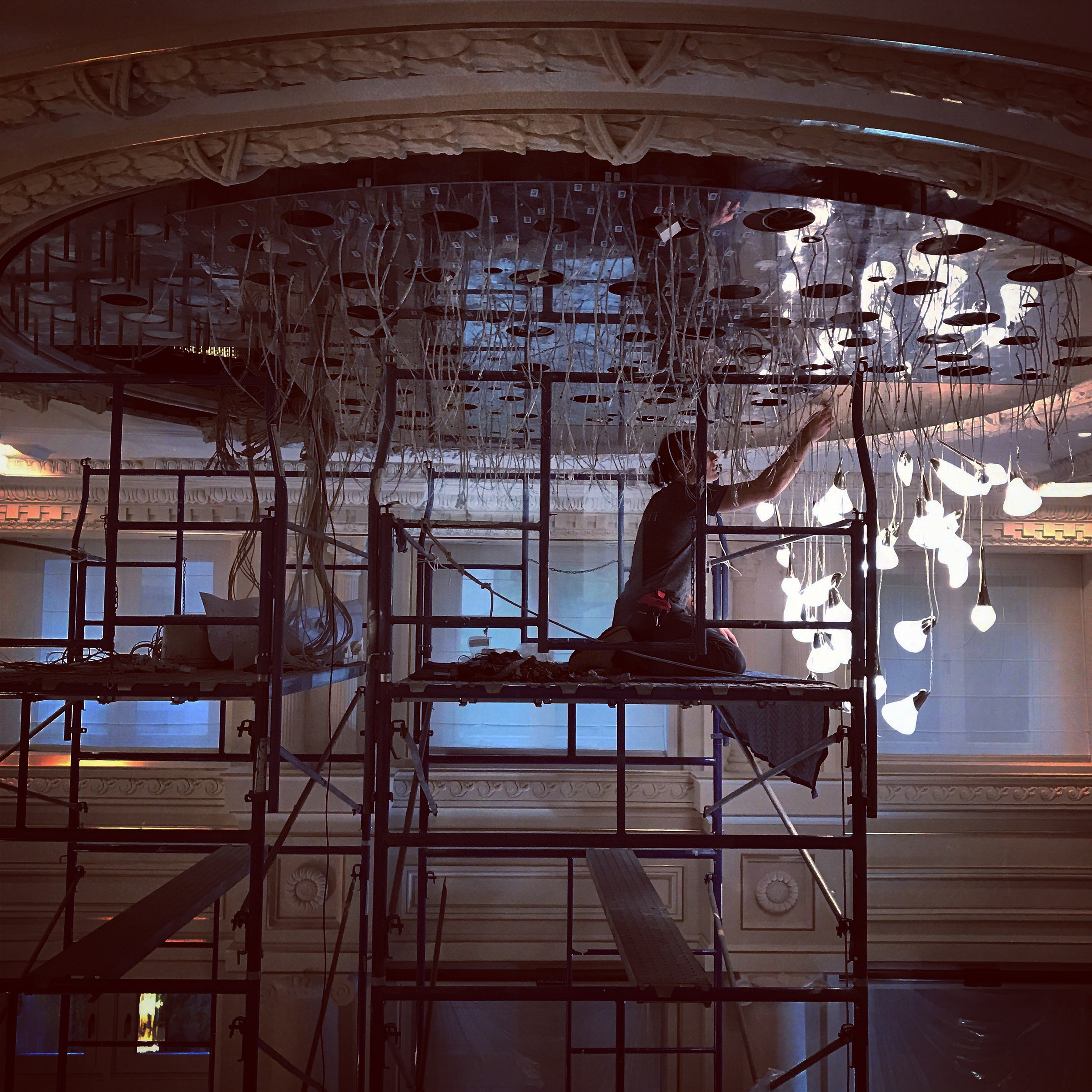 A worker on scaffolding installing or repairing a chandelier with multiple hanging lightbulb fixtures in an ornate, classical interior space.