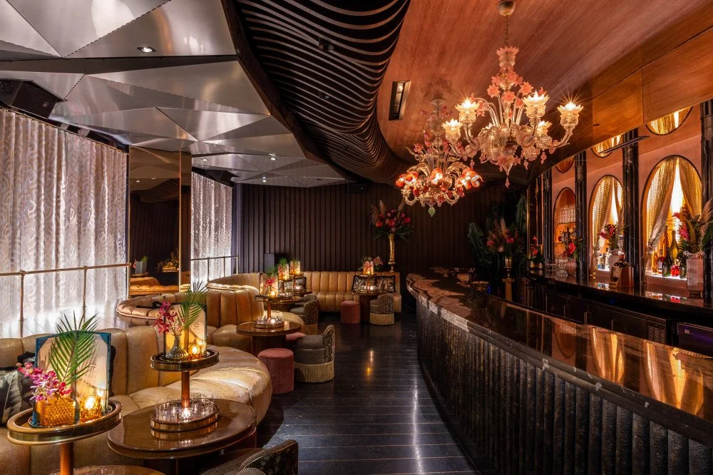 Elegant lounge with plush seating, small round tables with candles and flowers, a curved bar with a reflective surface, and elaborate chandeliers in a luxurious, dimly-lit atmosphere.