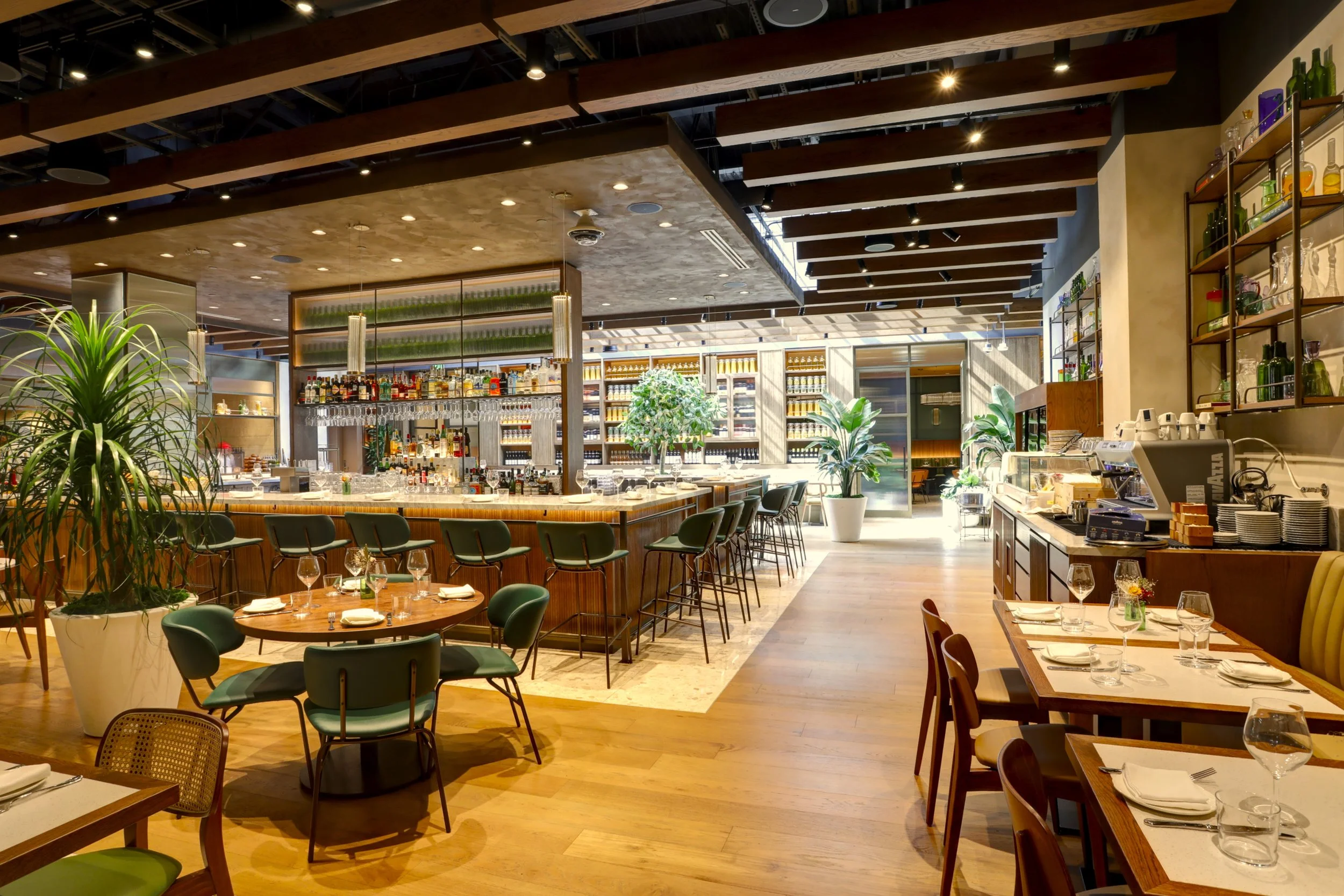 Inside a modern restaurant with a bar area, seating tables, and large plants, illuminated by ceiling lights and natural light.