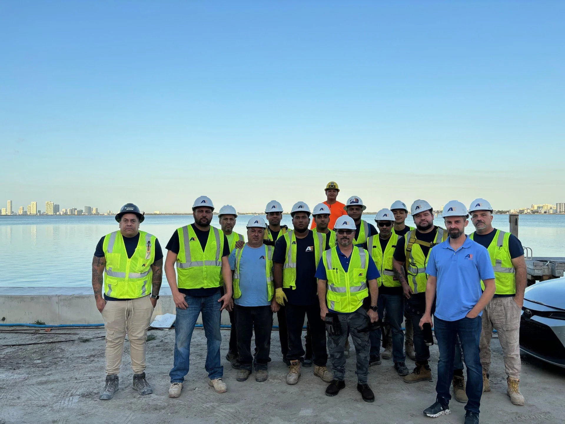 A group of construction workers and supervisors wearing safety vests and helmets at a waterfront construction site with cityscape in the background.