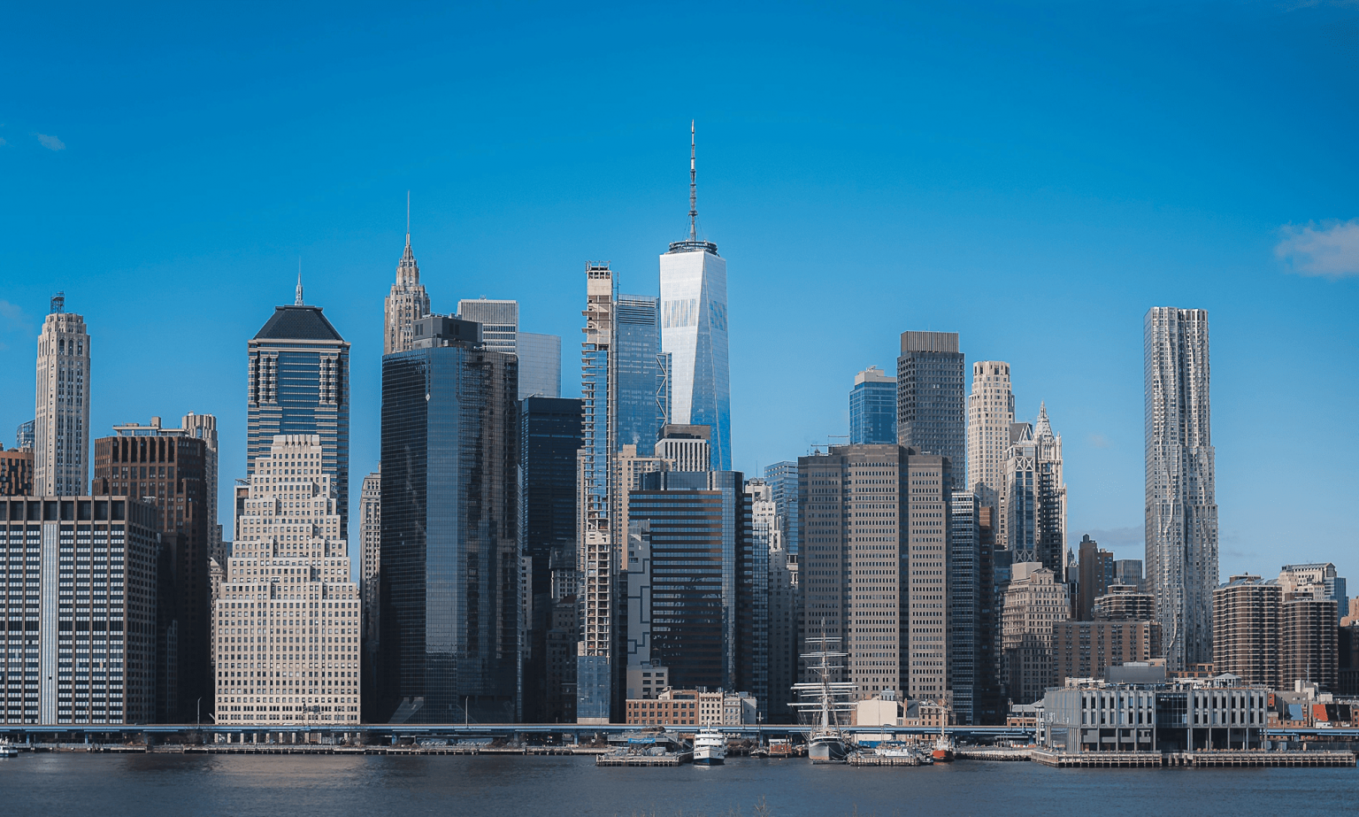Skyline of Manhattan, New York City with tall skyscrapers; One World Trade Center prominently in center, blue sky backdrop.