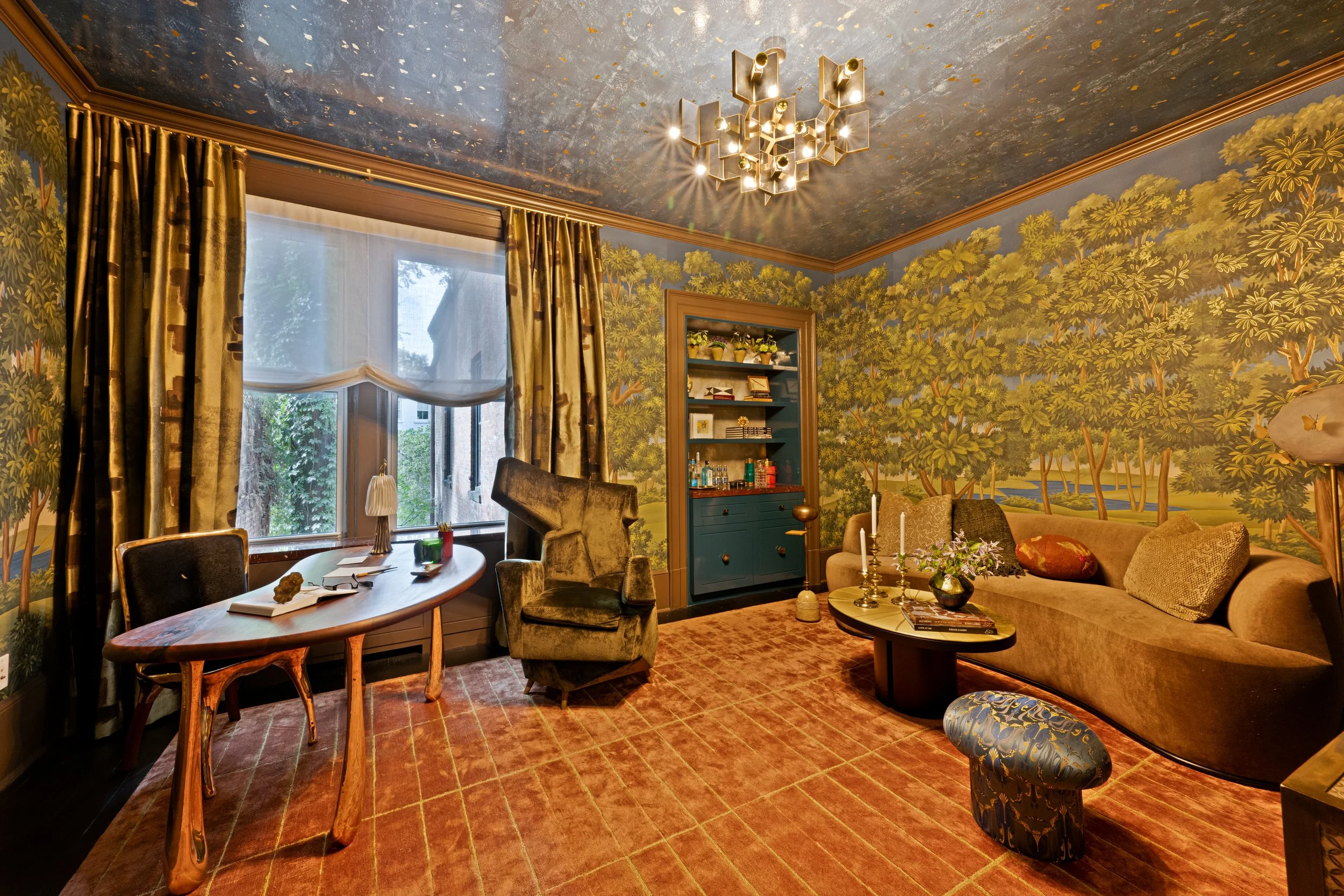 Living room with a mural of trees on walls, a chandelier, a window with curtains, a beige sofa with pillows, a coffee table with candles and flowers, a green armchair, a blue bar cabinet, and a patterned ottoman.