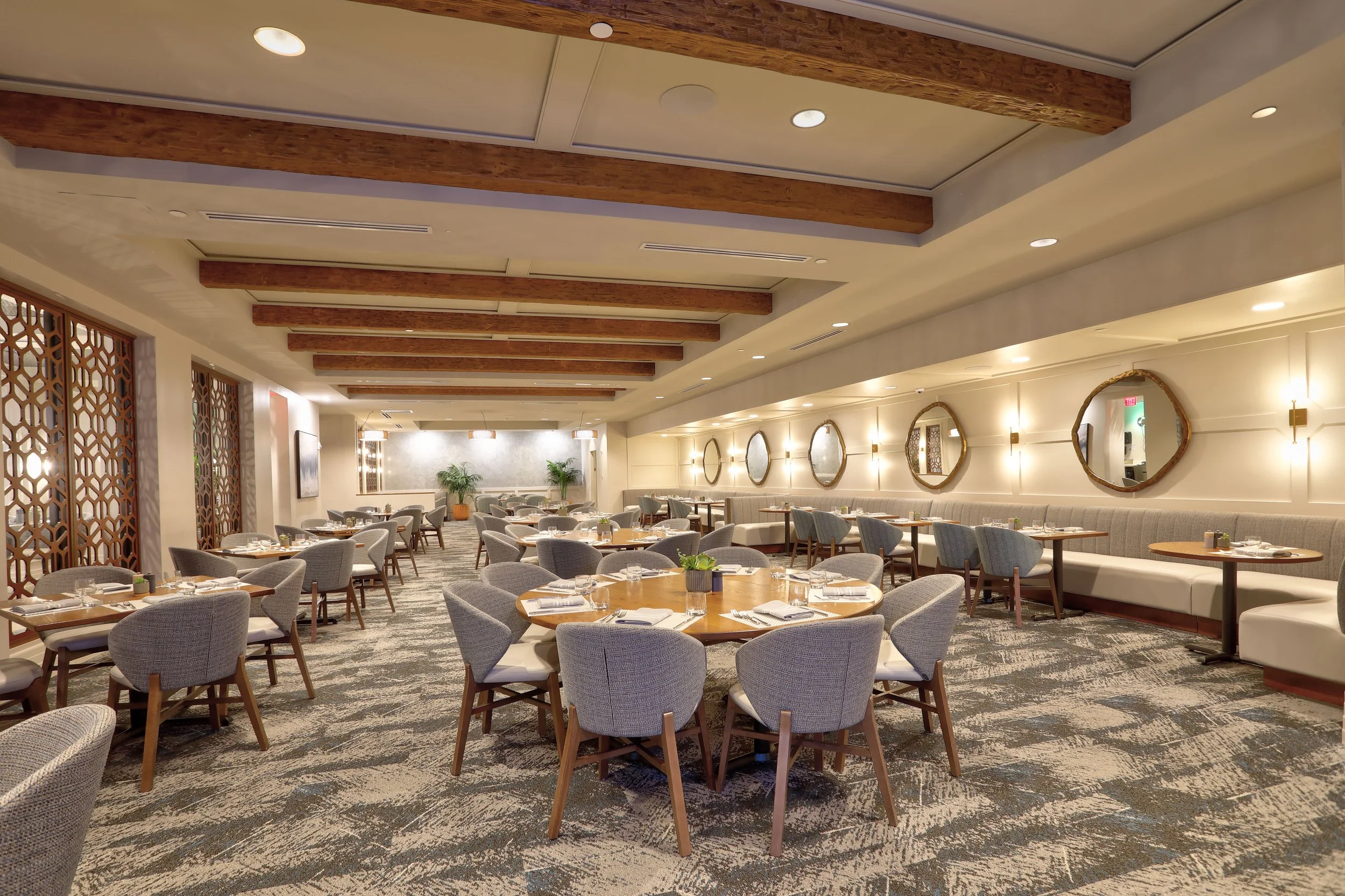 Elegant restaurant dining area with round tables set with silverware, glasses, and napkins, surrounded by upholstered chairs and a long white banquette along the wall. The room features wooden beams on the ceiling, circular mirrors on the wall, and potted plants at the back.