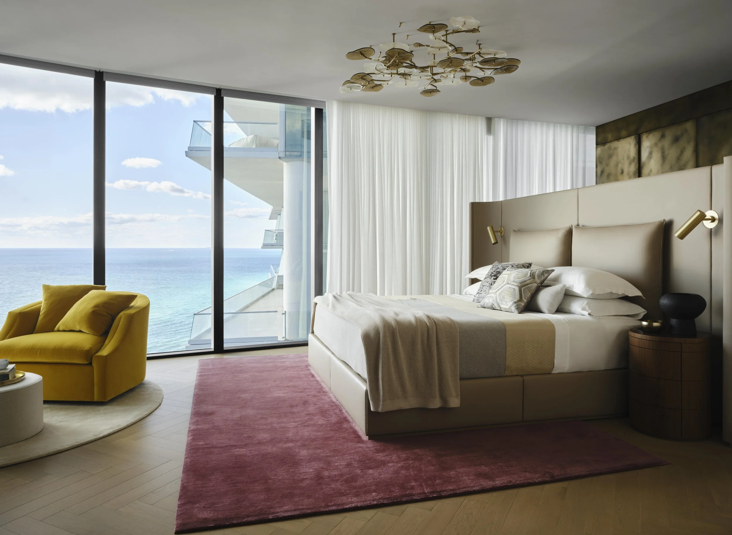 Luxury hotel room with a large bed, a yellow armchair, and a view of the ocean through floor-to-ceiling windows.