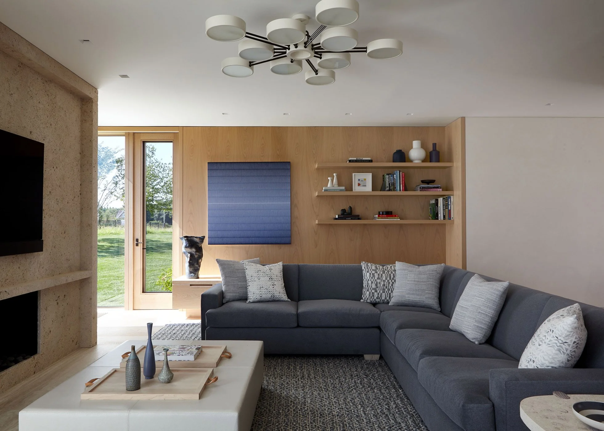 Modern living room with a gray sectional sofa, a white ottoman with decorative vases, a built-in wooden shelf with books and vases, a large abstract blue wall art, a window showing greenery outside, and a ceiling light fixture.