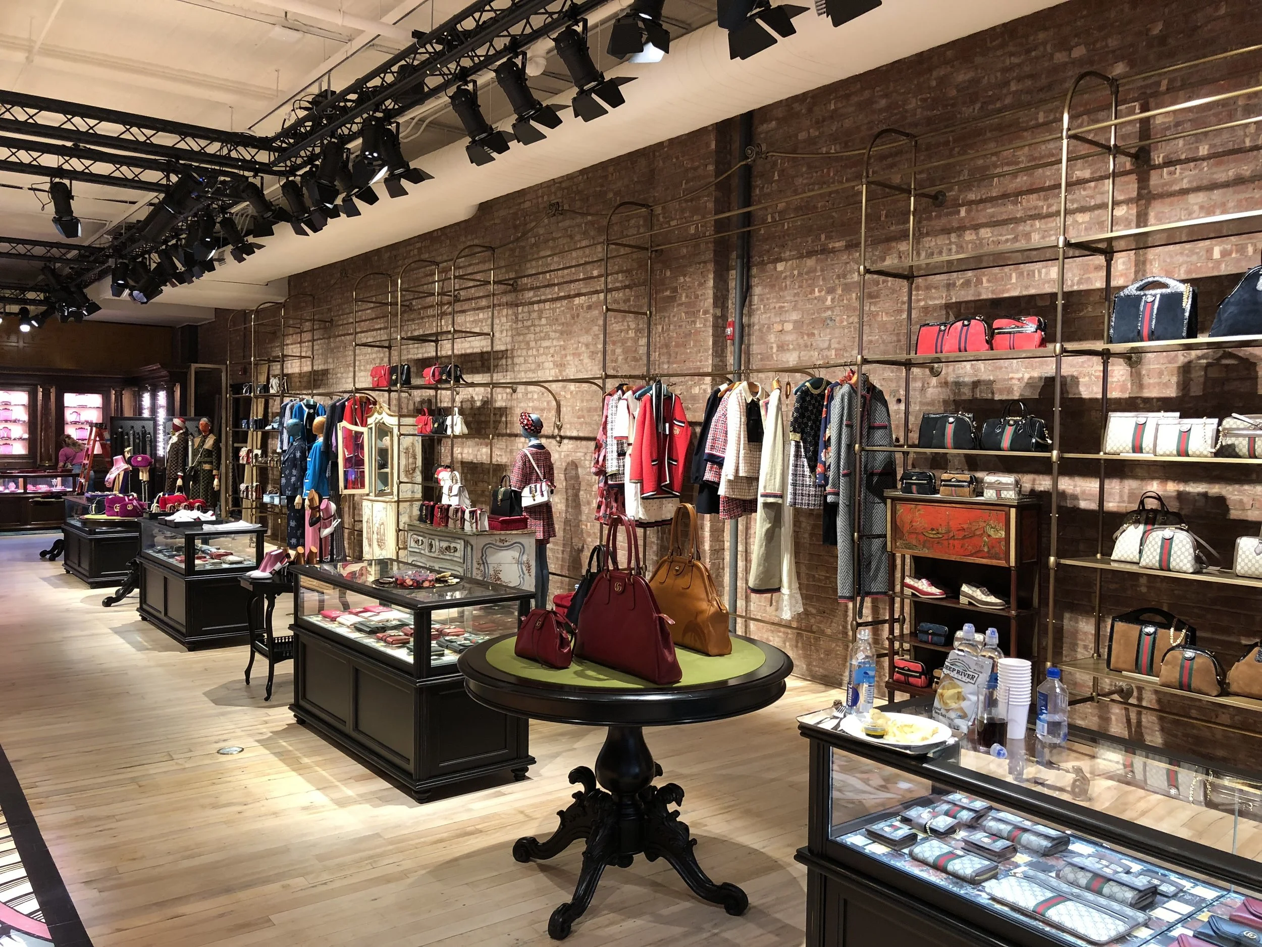 Interior view of a boutique store with shelves of handbags, mannequins dressed in clothing, and glass display cases showcasing accessories on a wooden floor, brick walls, and ceiling-mounted lighting.