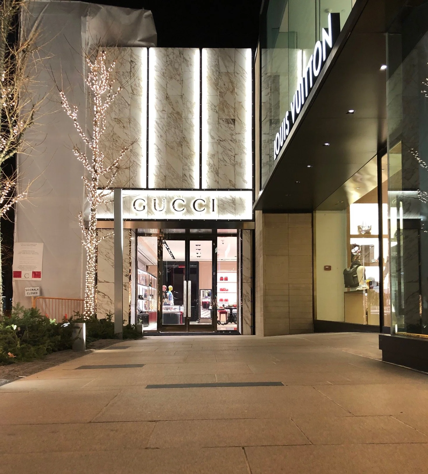 Night view of a luxury storefront with Gucci and Louis Vuitton signage, glass windows displaying products, and decorative lighting on a tree outside.