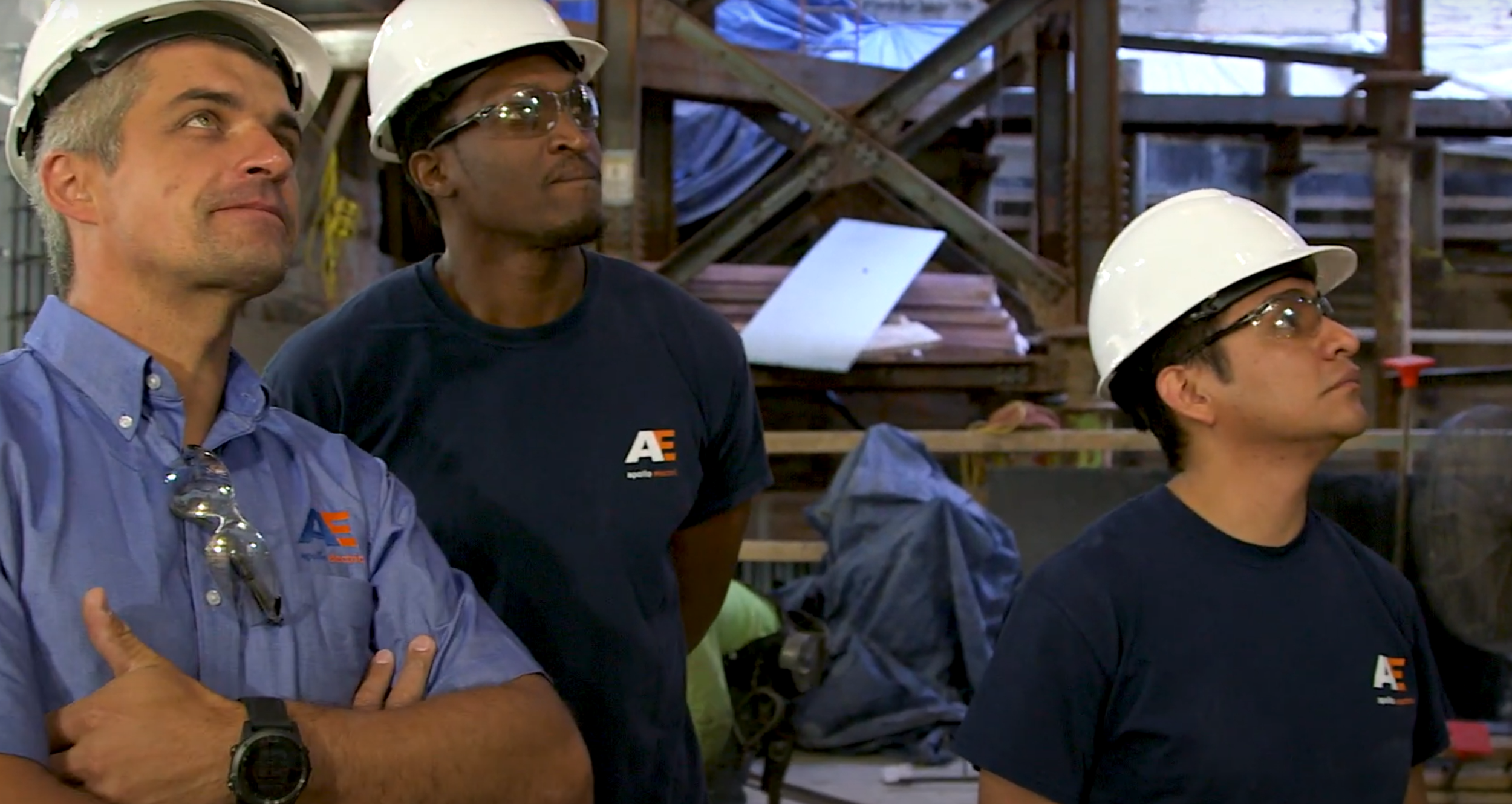 Three construction workers in hard hats and safety glasses in a workshop or construction site, standing attentively and listening.