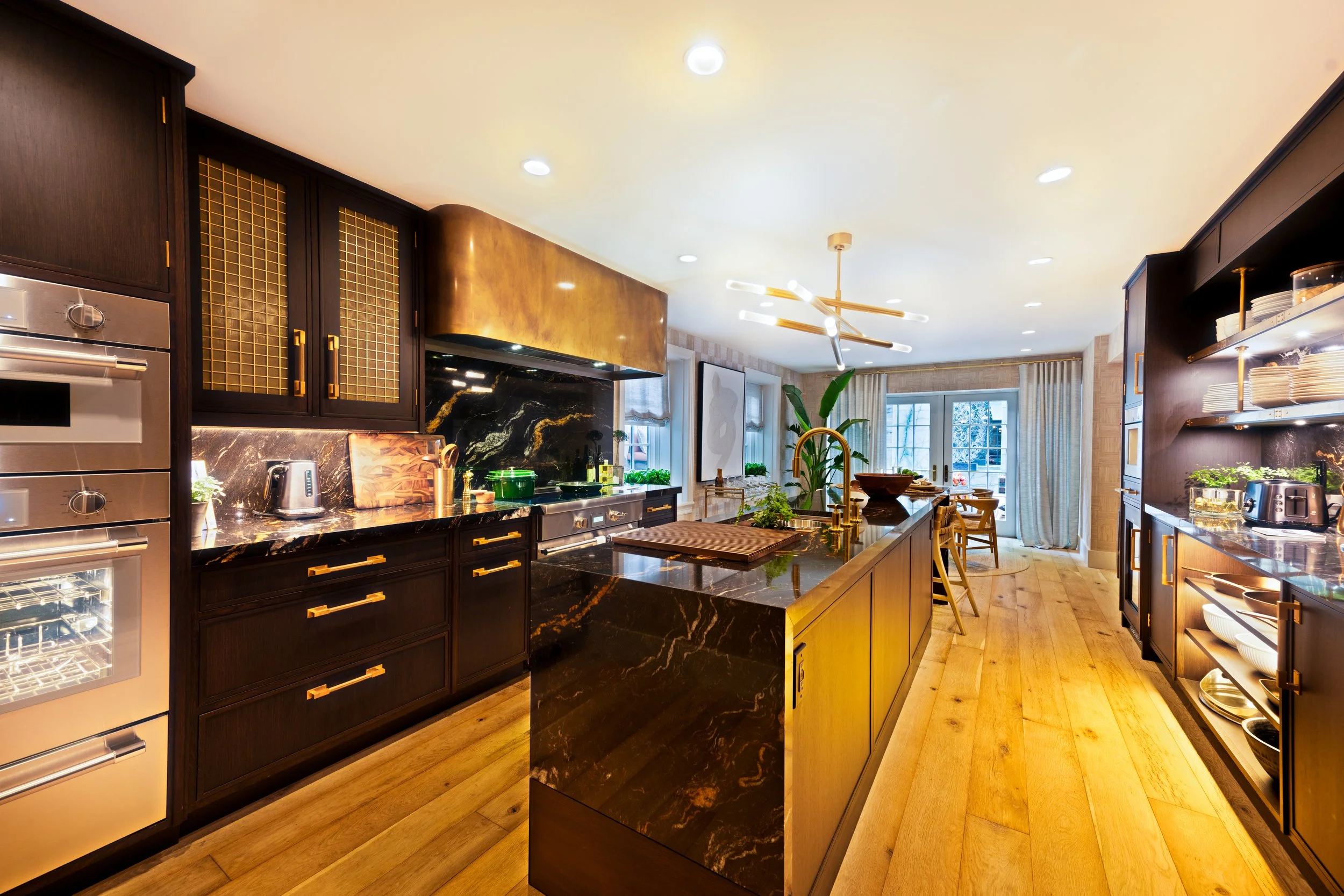 Modern kitchen with dark cabinetry, marble countertops, and wooden flooring, illuminated by ceiling lights and adorned with plants and artwork.