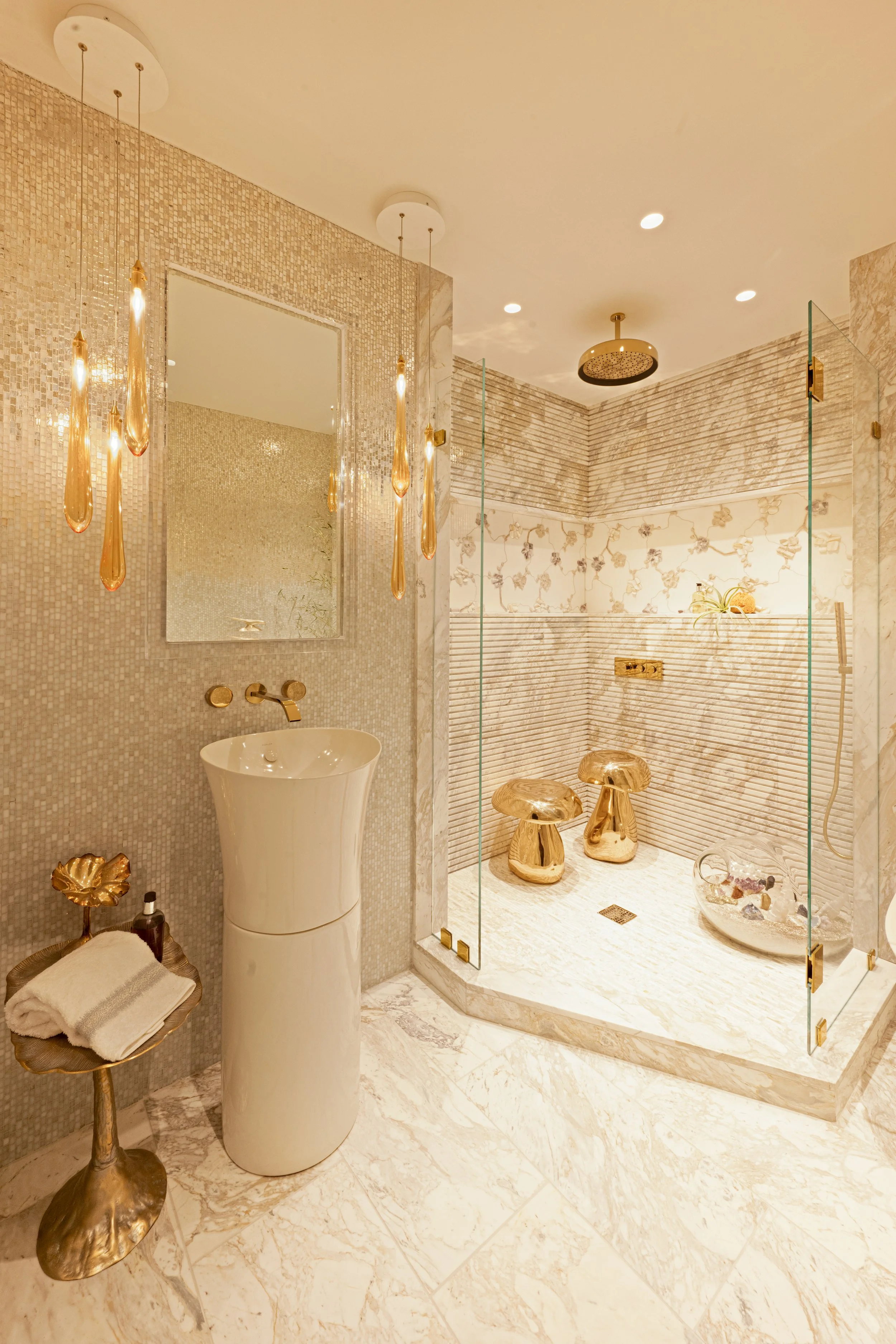 Luxurious bathroom with gold accents, a pedestal sink, a mirror, a mosaic tiled wall, and a glass-enclosed shower with mushroom-shaped stools inside.