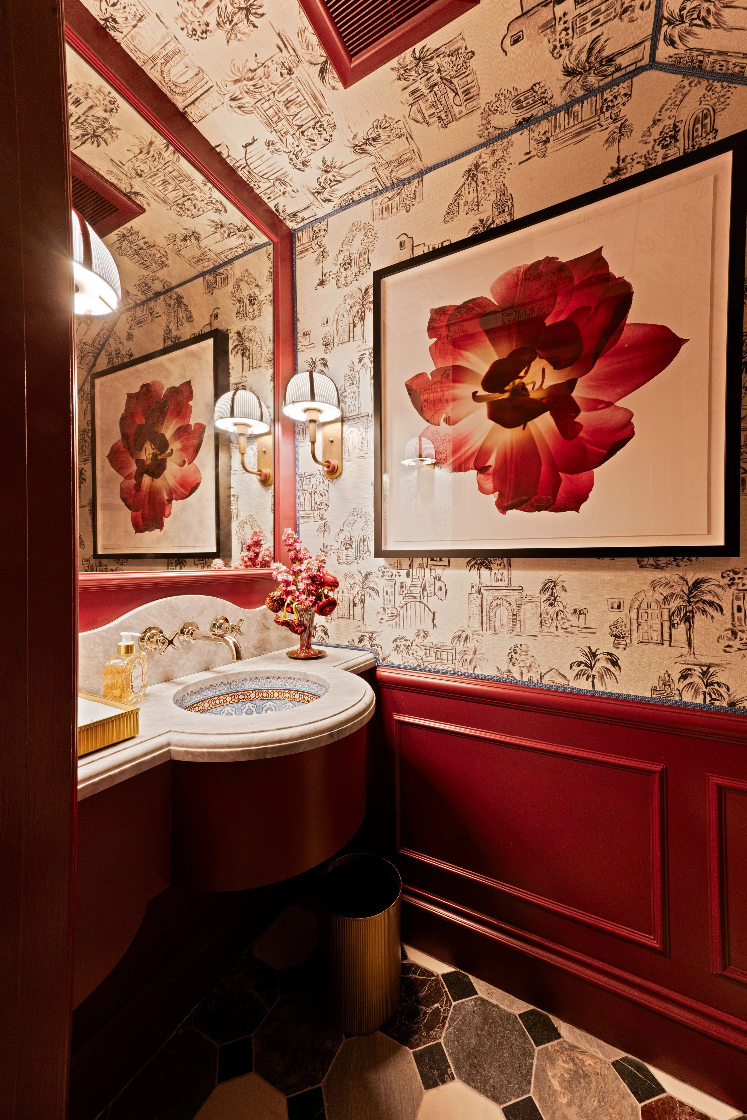 A small bathroom with a red and white color scheme, featuring a large floral artwork on the wall, a mirror, wall sconces, a marble sink, a vase of pink flowers, and patterned wallpaper with architecture and palm tree motifs.