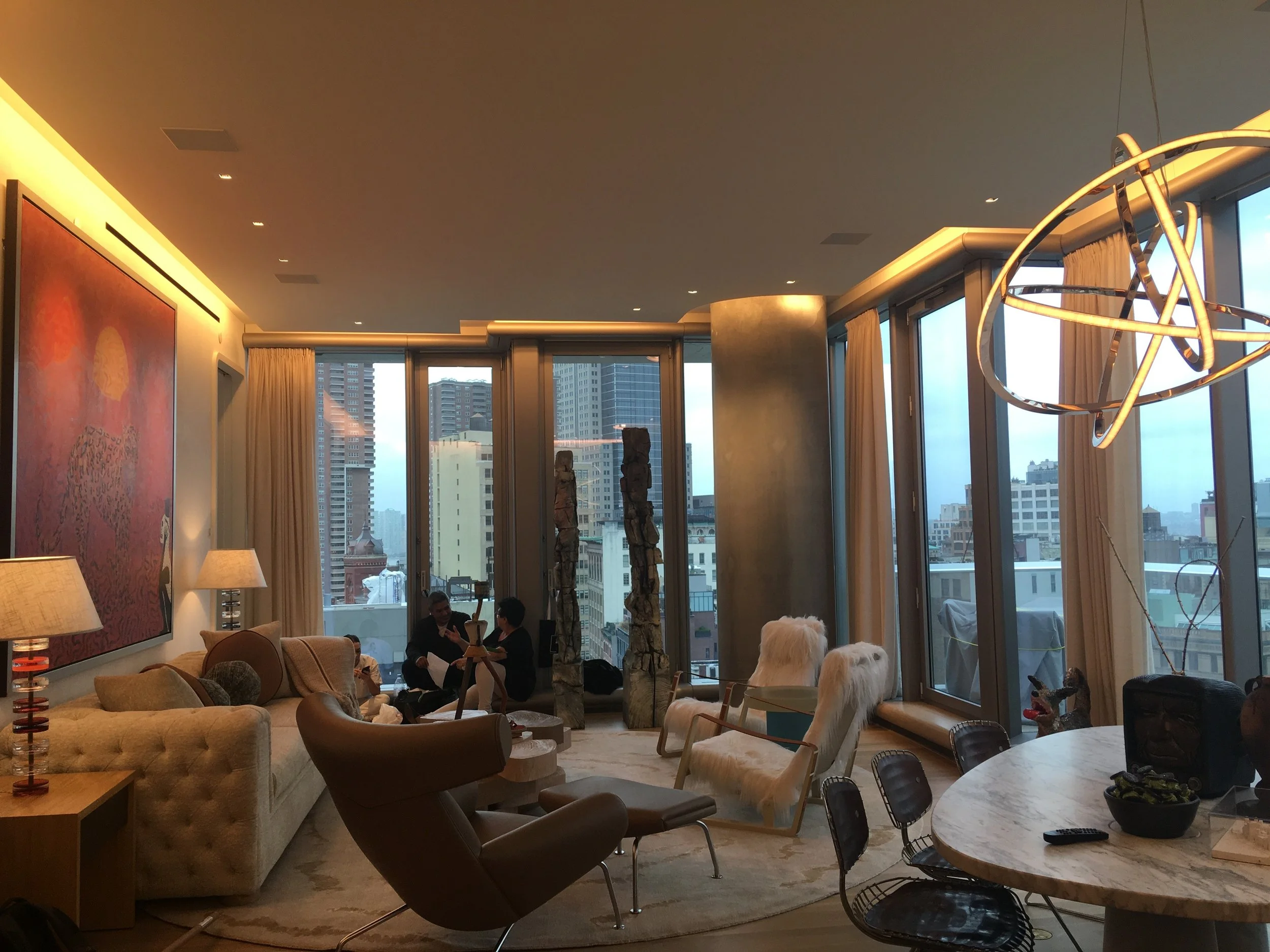 Modern living room with large windows showing city skyscrapers, beige sofas, armchairs, side lamps, abstract artwork on the wall, decorative sculptures, and a round marble table with chairs.