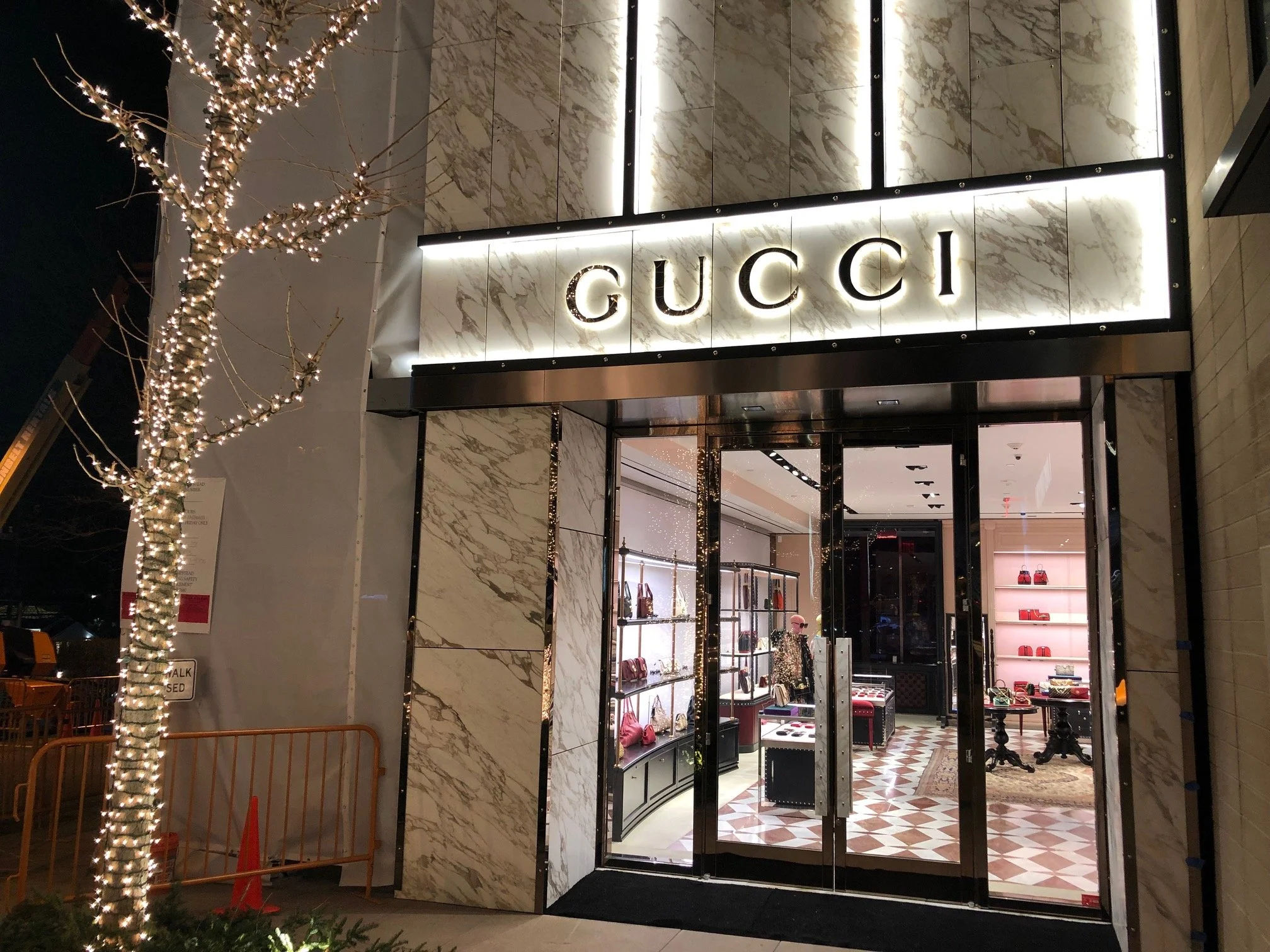 Night view of a Gucci storefront with illuminated sign, glass doors, decorated with lights on a tree outside, and handbags displayed inside.