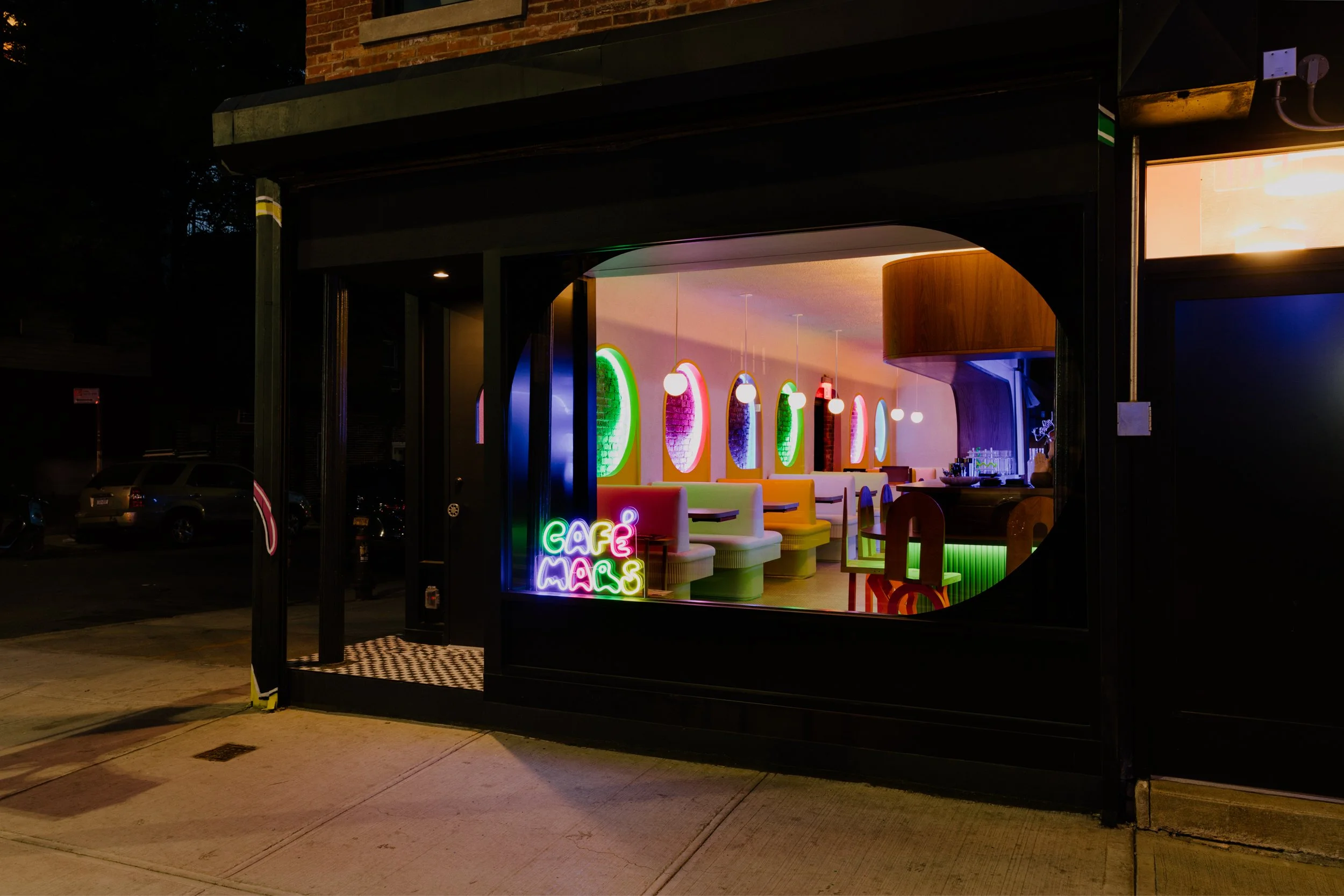 Colorful interior of a cafe with neon signs, booths, and round windows, seen through a large window at night.