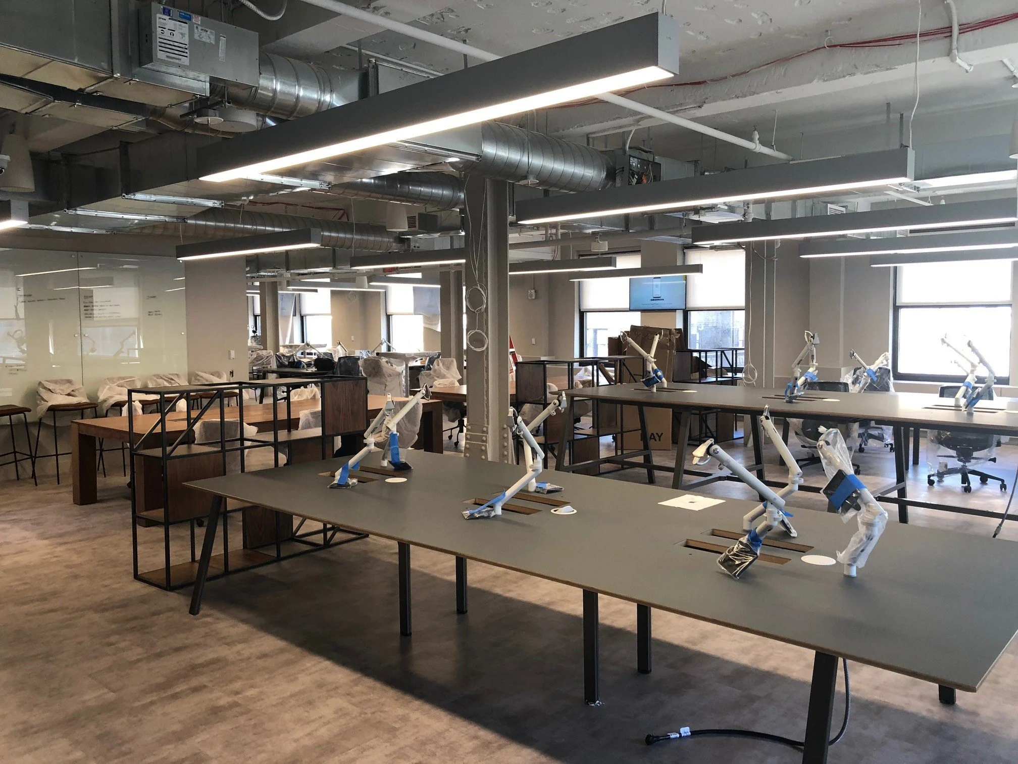 Empty workspace with several robotic arms on tables, some boxes and chairs in the background, large windows, industrial ceiling with exposed ducts, and a polished wooden floor.