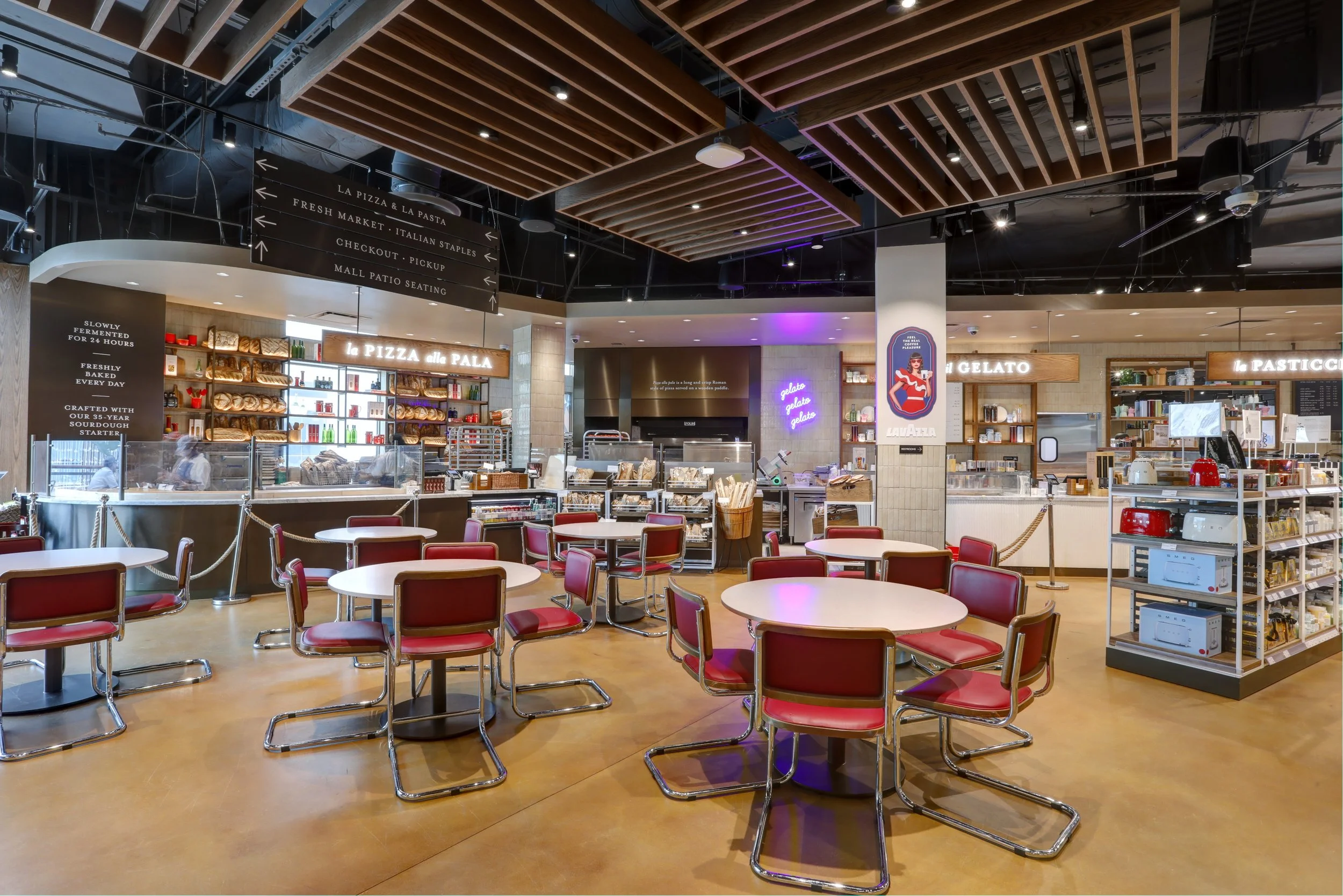 Inside a modern restaurant or cafe with red-seat tables, bakery shelves, and signage for pizza, gelato, and pastries, with a black ceiling and wooden accents.