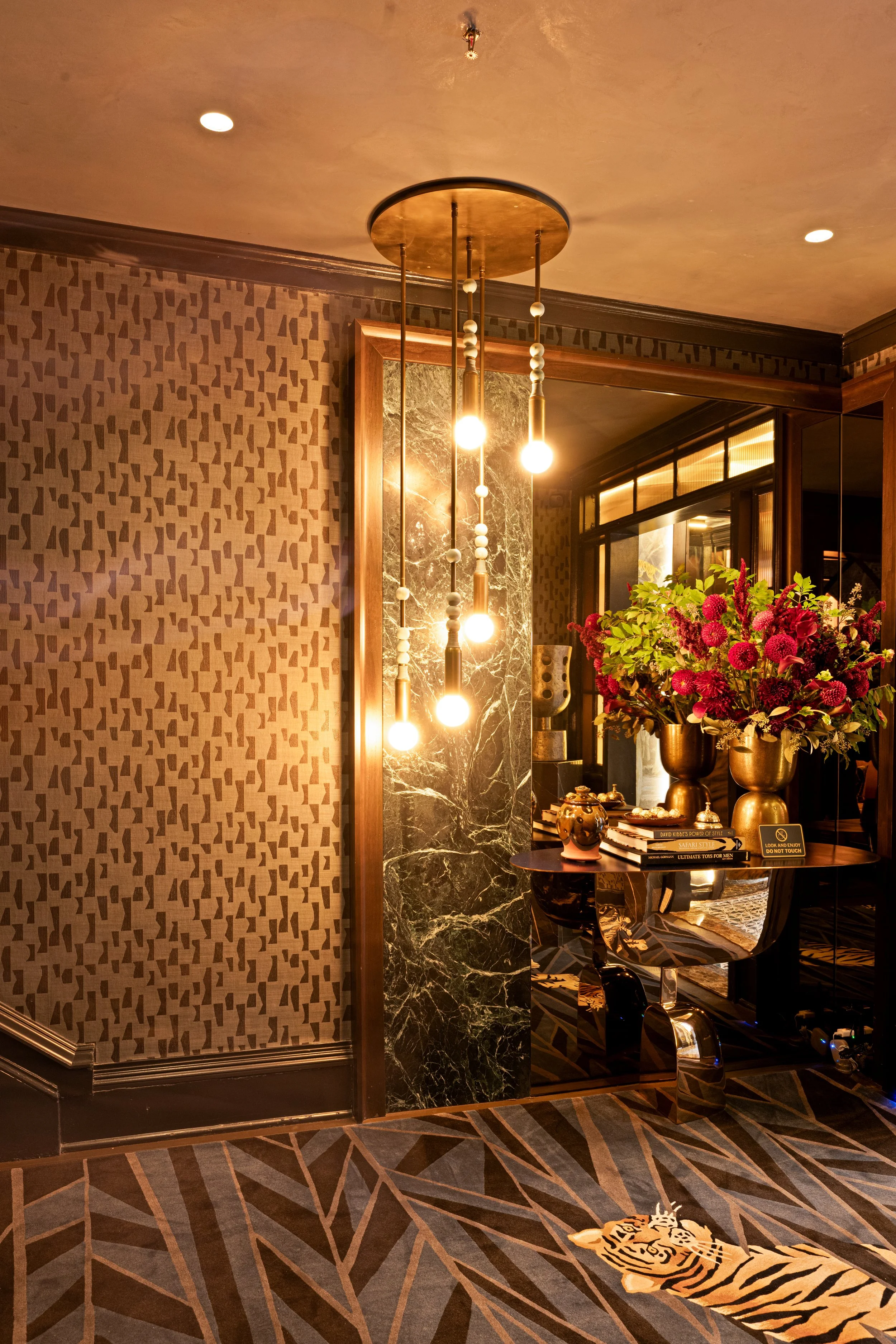 Interior of a luxurious hotel lobby with a modern chandelier, a table with flower arrangements, and decorative details.