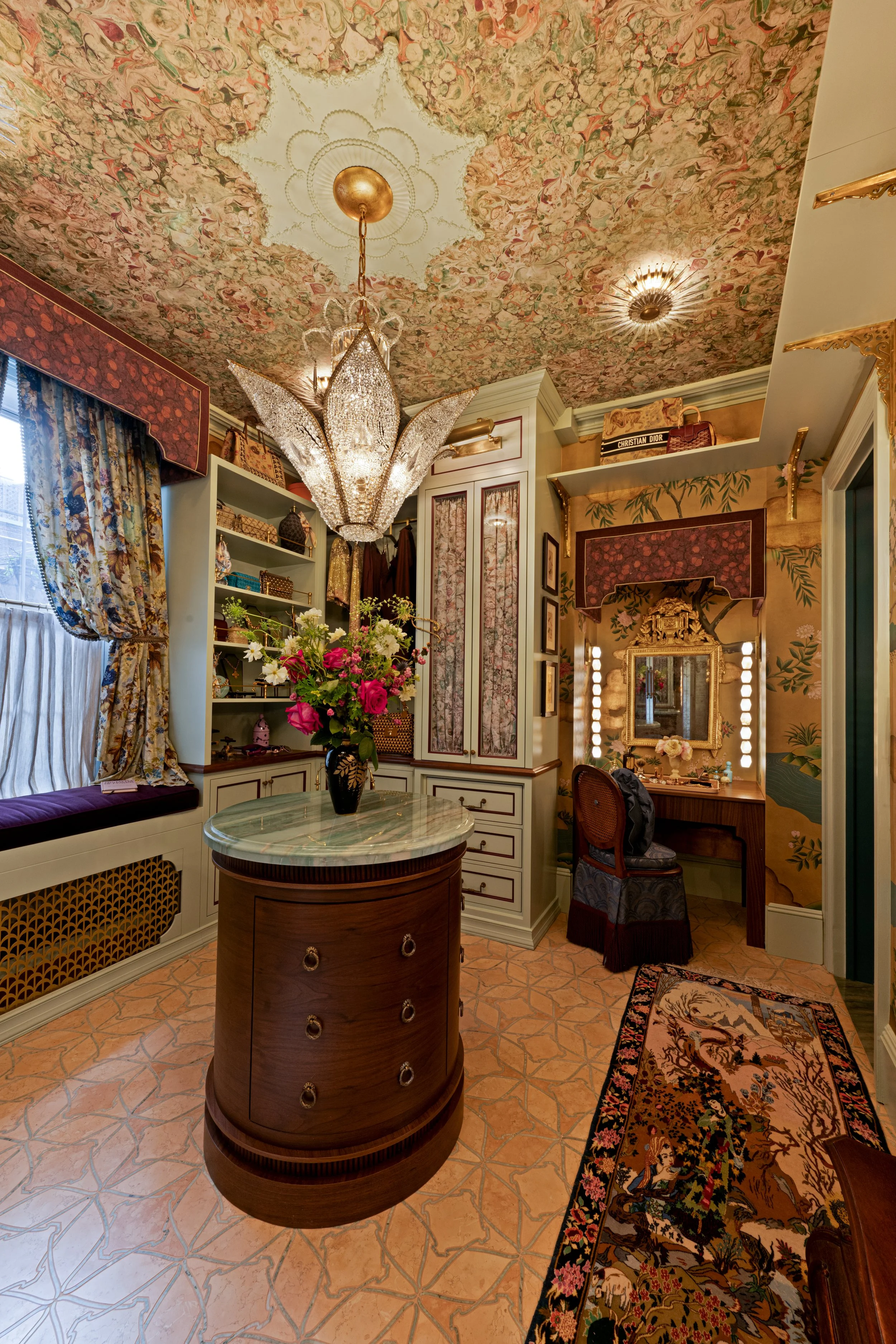 A luxurious room with floral wallpaper, a chandelier, a vase of pink and white flowers on a marble-topped cabinet, a window with floral curtains and a cushioned window seat, and a vanity mirror with lights, all decorated in a vintage style.