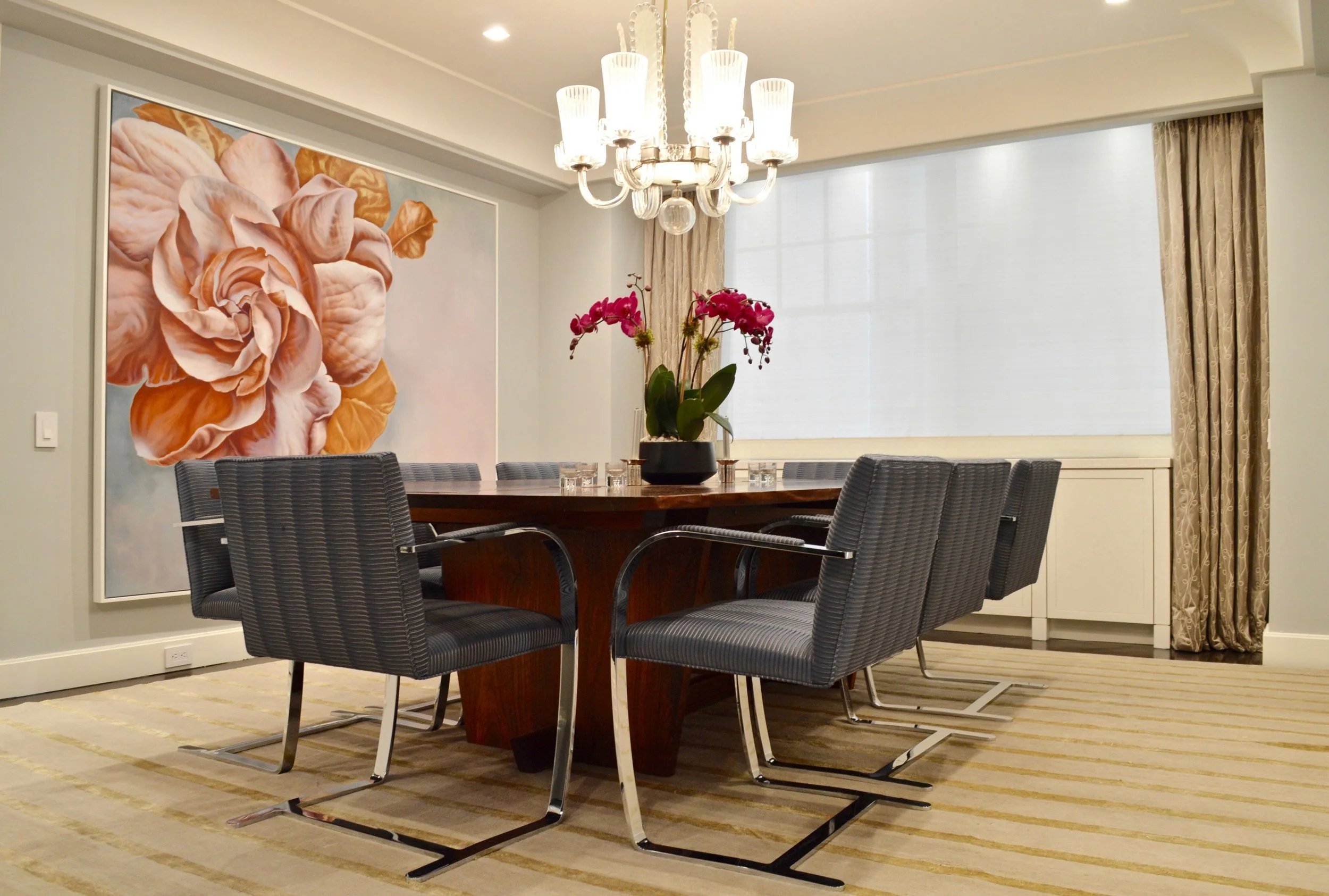A formal dining room with a large wooden round table surrounded by eight black striped chairs. The table has a centerpiece of pink orchids in a black vase. A chandelier hanging from the ceiling illuminates the room. There is a large floral painting on the wall to the left, and a window with cream-colored curtains on the right. The floor is covered with a beige striped carpet.