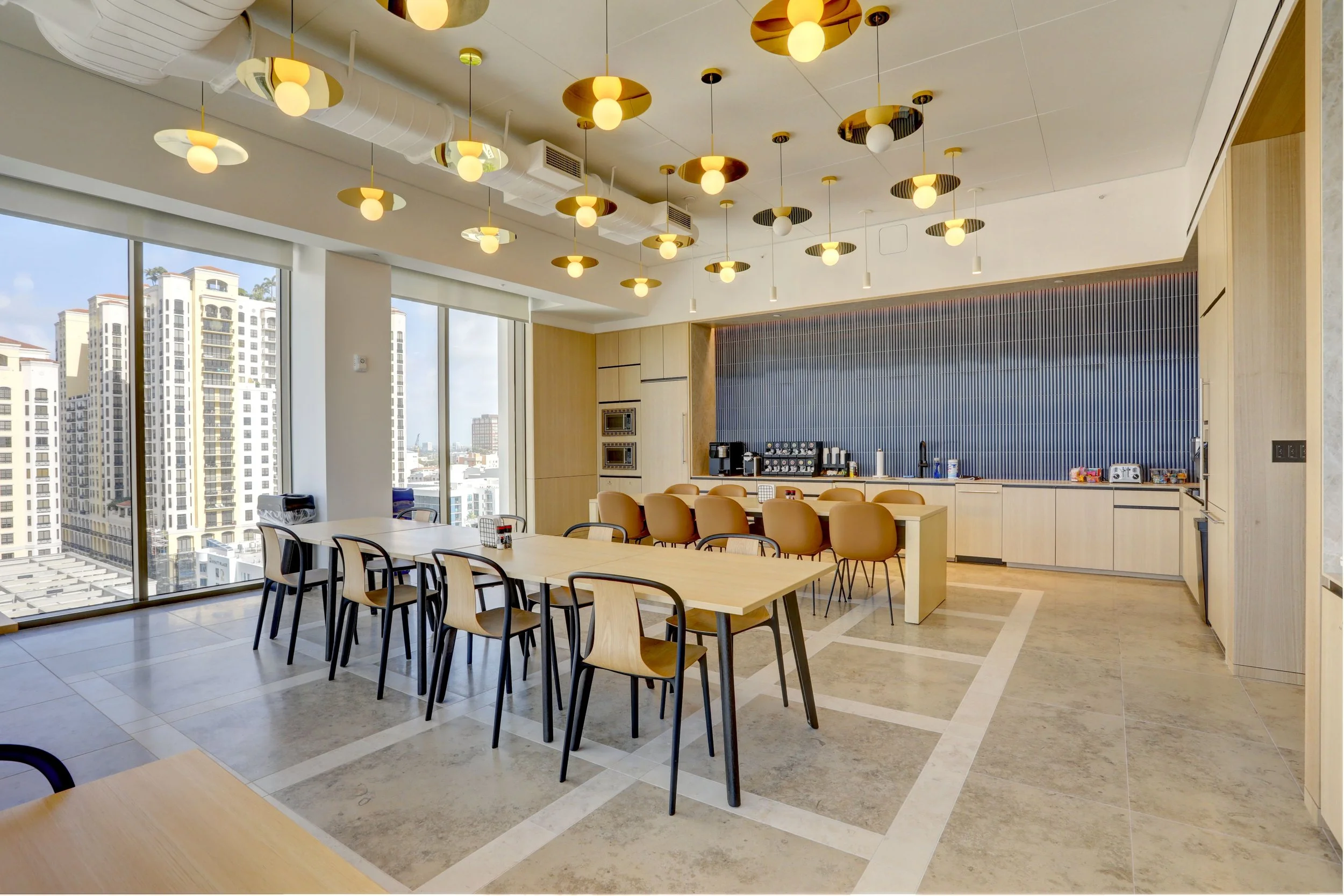 Modern office breakroom with large windows showing a city skyline, with a table, chairs, and a coffee station.