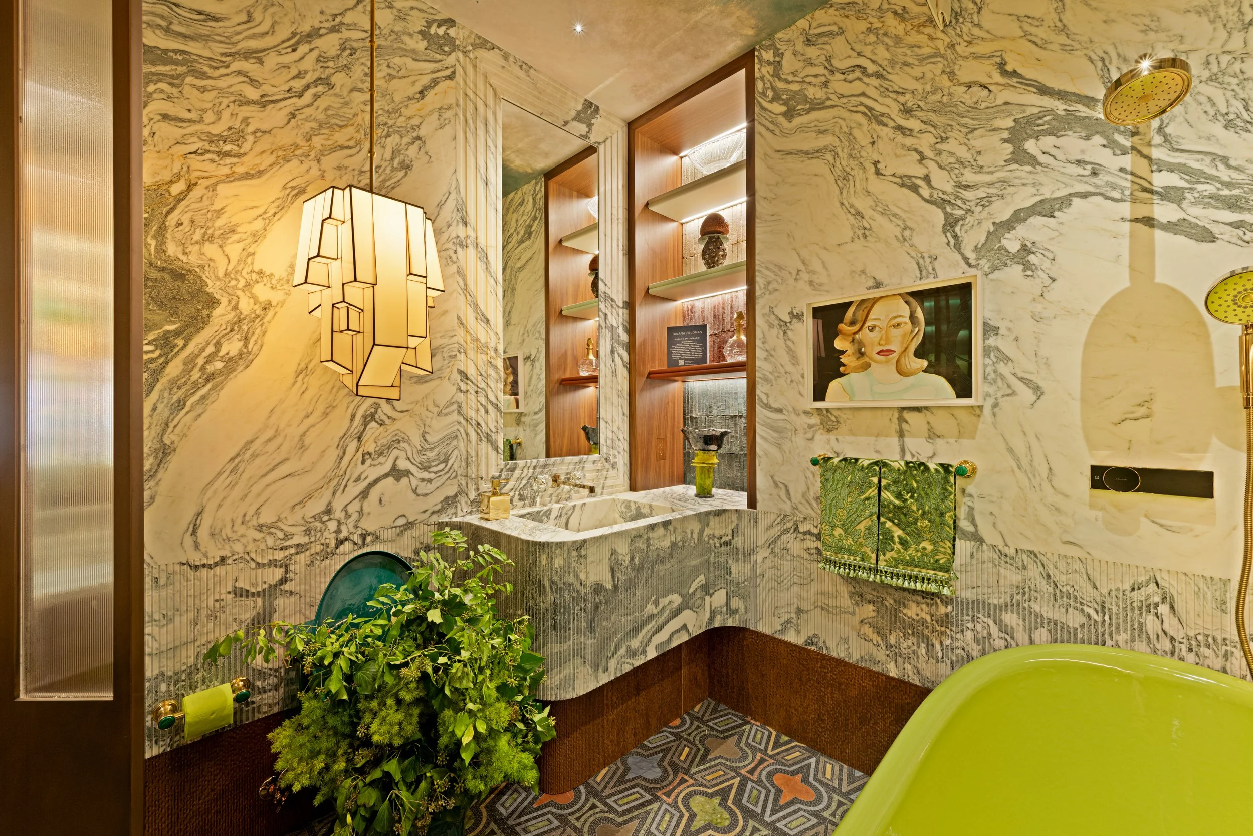 A luxurious bathroom with marble walls and a marble sink. There is a large mirror, a green towel, a framed artwork of a woman, plants, and unique lighting fixtures. The floor features geometric patterned tiles and a yellow showerhead is visible.