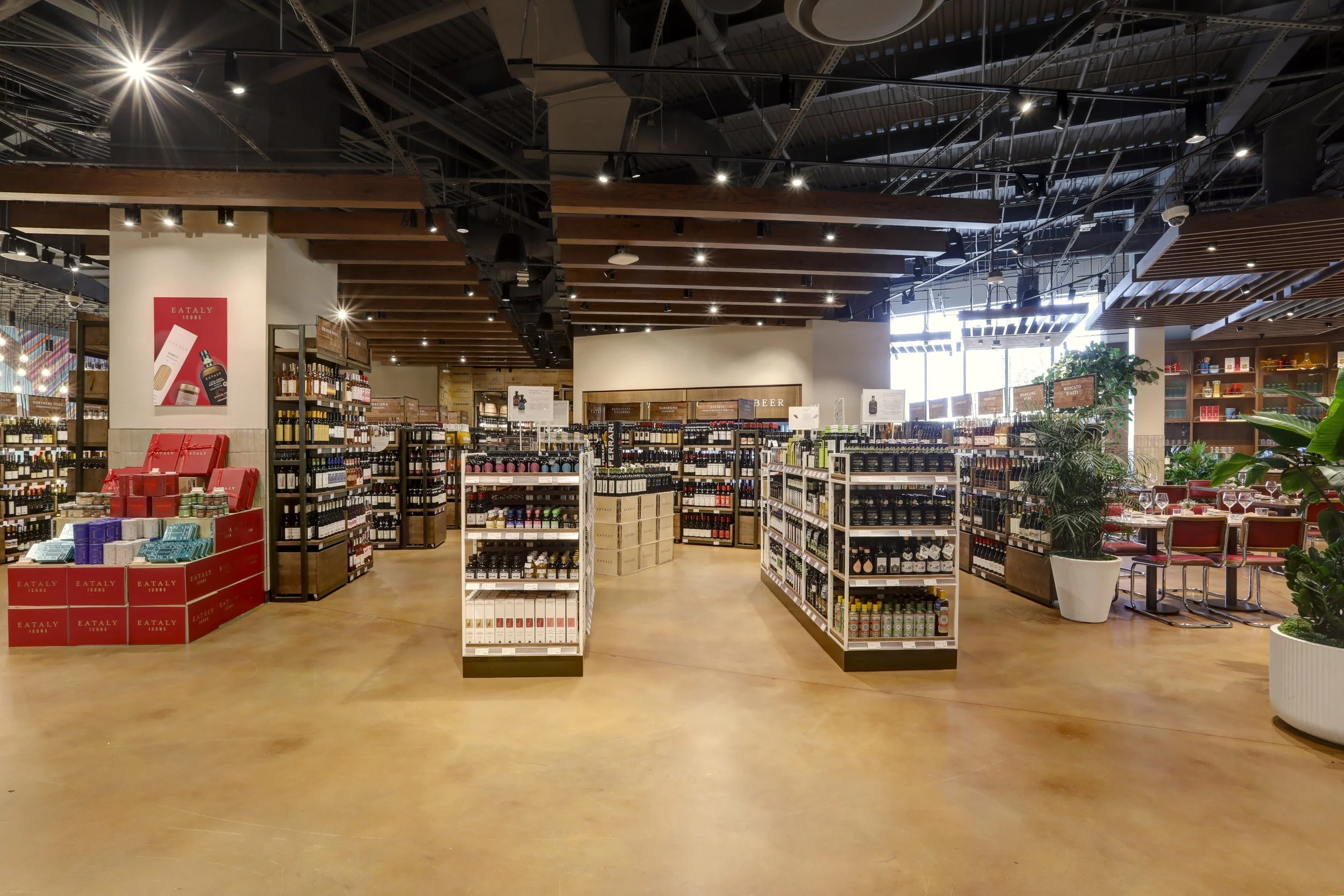 Interior of a retail store with shelves stocked with wines, a display with gift boxes on the left, and a dining area with tables and chairs on the right, decorated with plants.