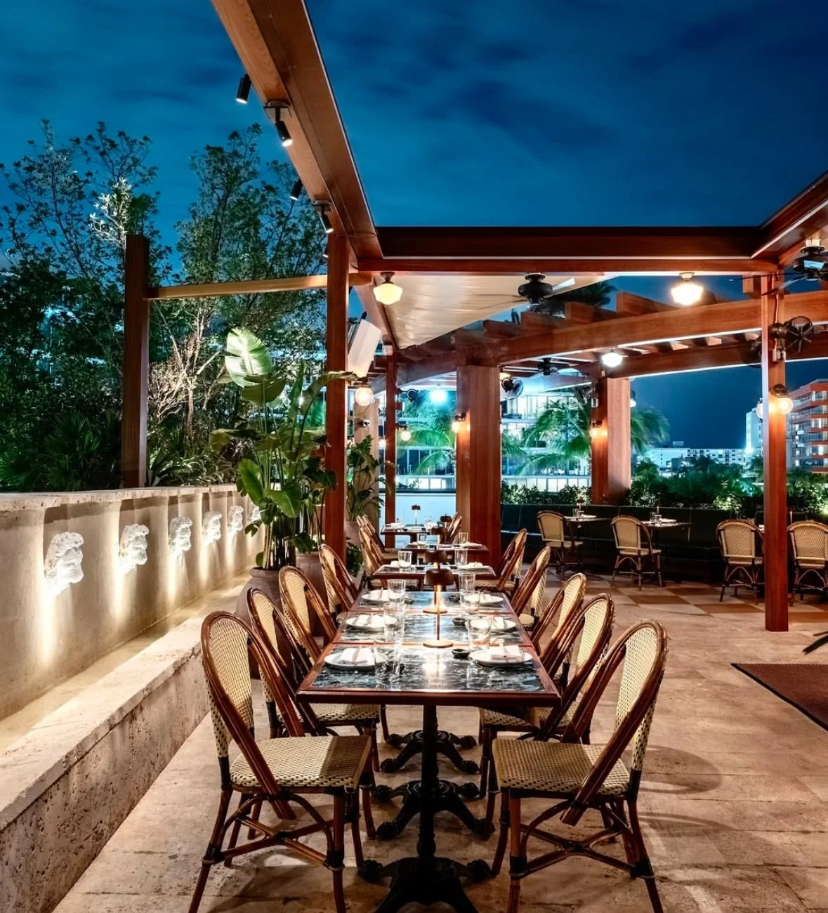 Outdoor rooftop restaurant at night with a long dining table, chairs, lush greenery, and city skyline in the background.