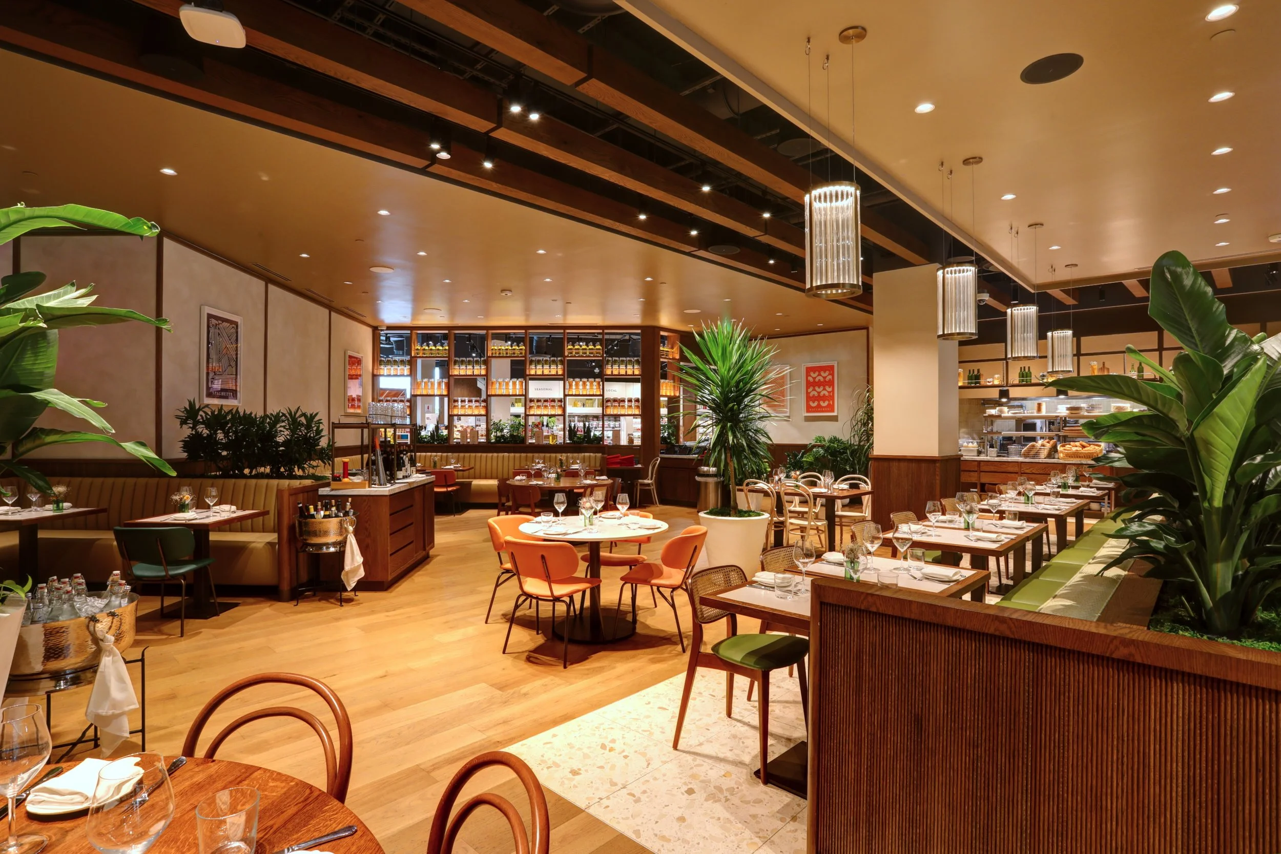 Interior of a restaurant with wooden flooring, green plants, and a bar area in the background.