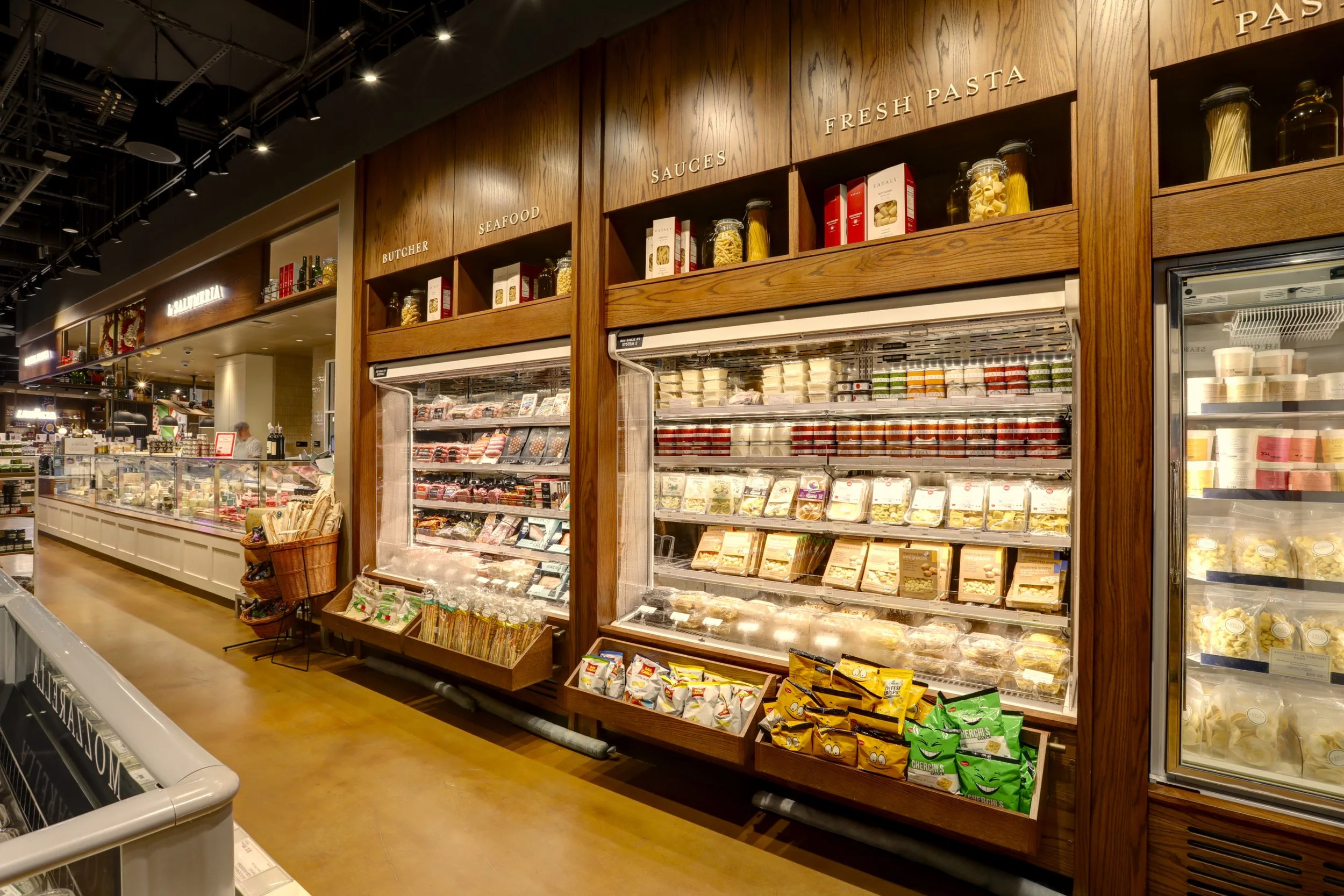 Grocery store aisle with refrigerated section displaying pre-packaged pasta, sauces, and dairy products, with wooden shelving and signs labeled 'Seafood', 'Sauces', and 'Fresh Pasta'.