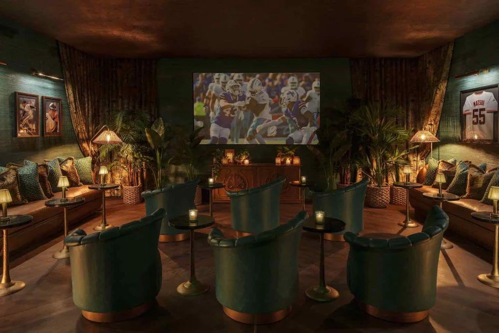 A cozy living room decorated with football memorabilia, featuring a large screen showing a football game, surrounded by plush chairs, sofas, and plants with dim lighting and small lamps.