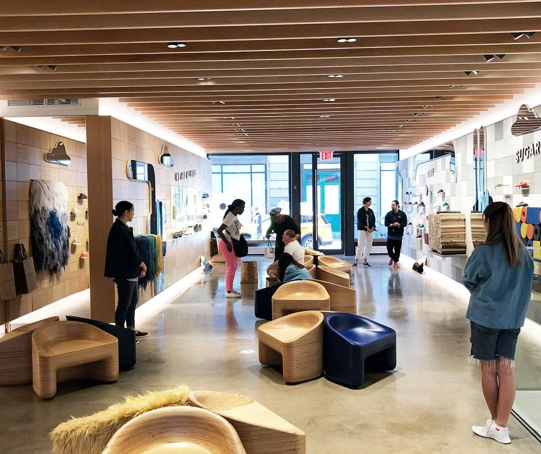 People shopping inside a store with wooden and colorful display shelves and benches, glass door entrance, and a wooden ceiling.