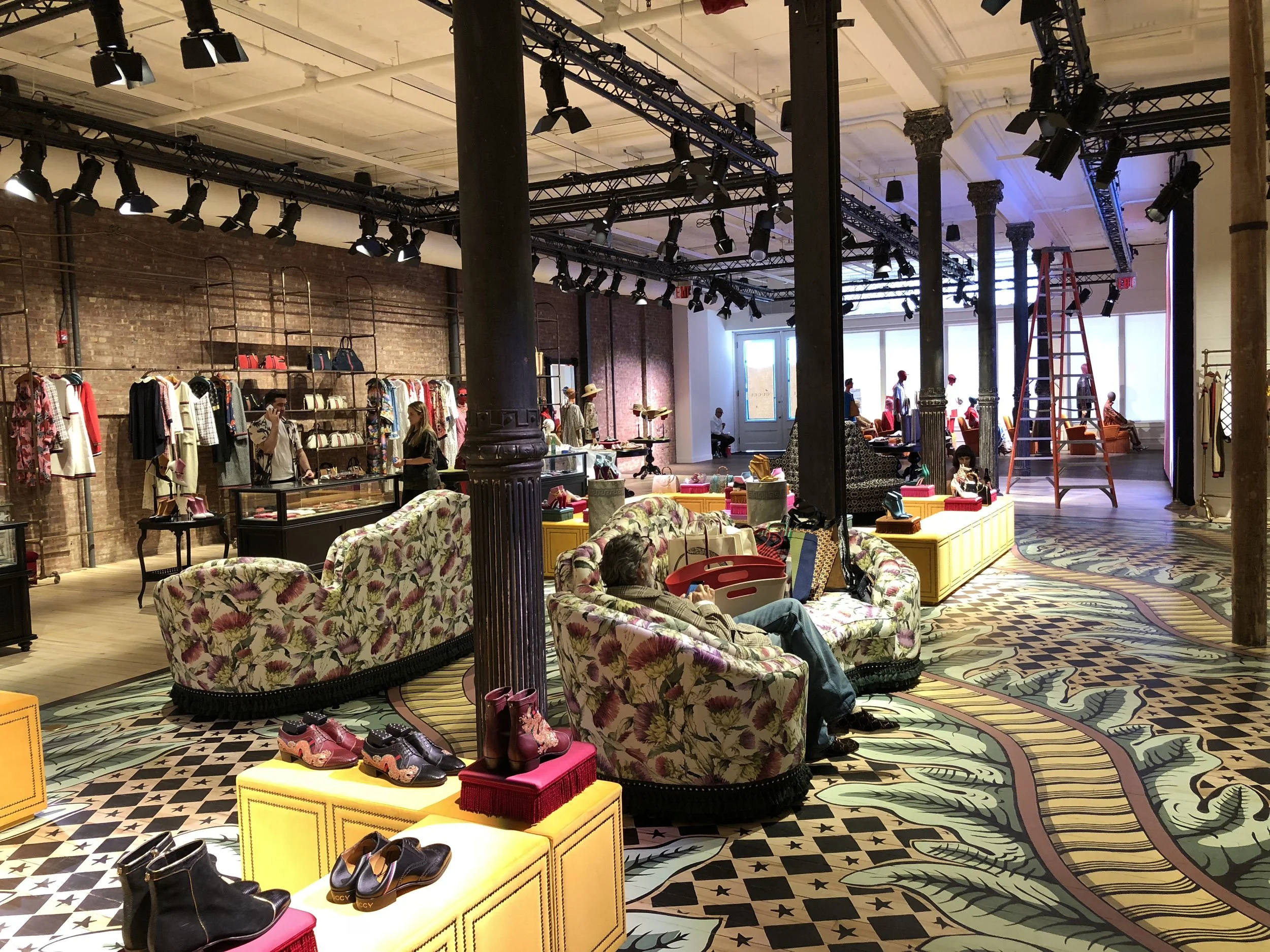 Interior of a retail store with vintage decor, floral sofas, clothing racks, and displays of shoes and handbags.