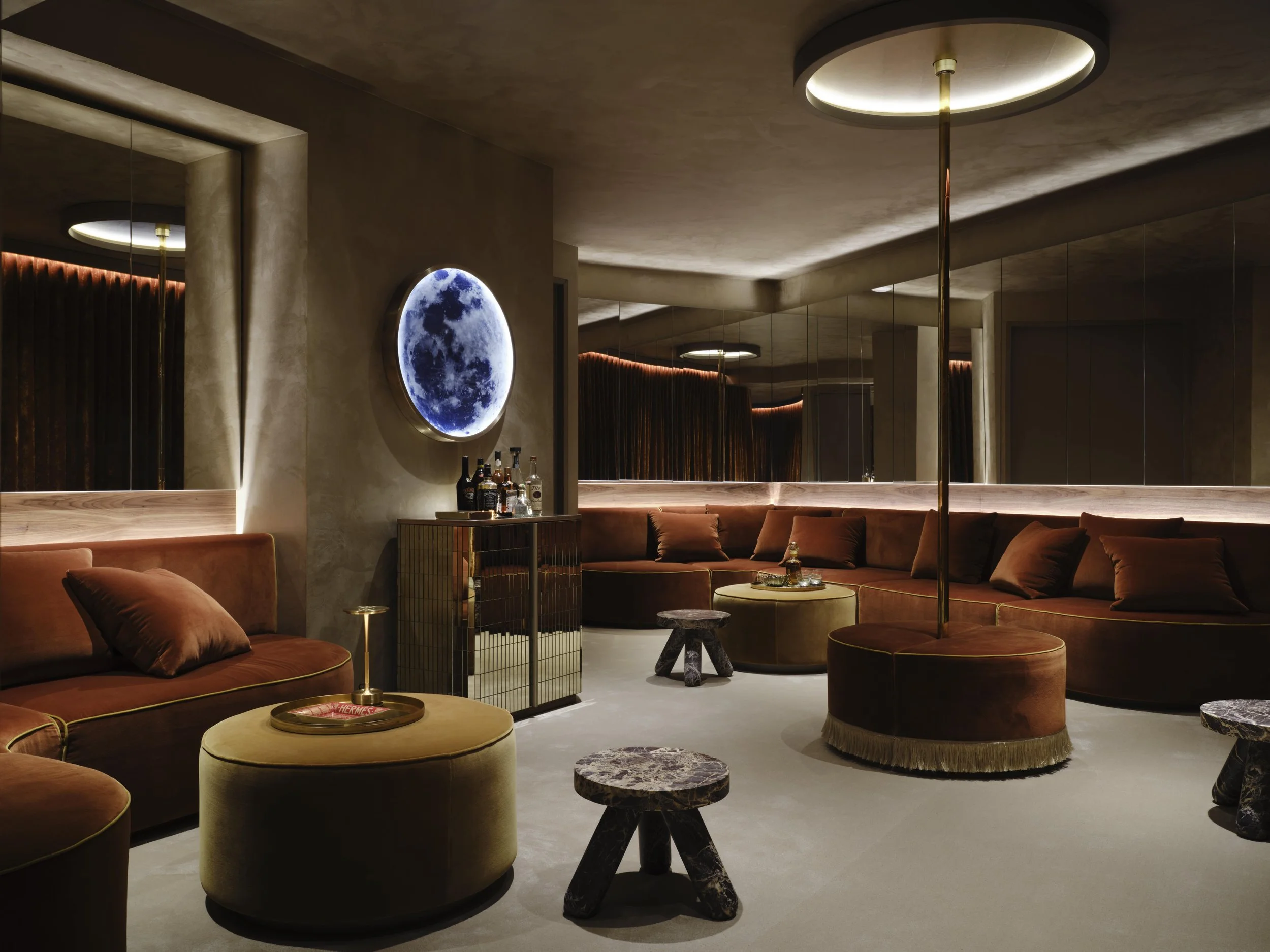 Luxurious modern lounge with brown velvet sofas, circular ottomans, small marble-topped tables, a bar cart, a moon photo on the wall, and dim ambient lighting.